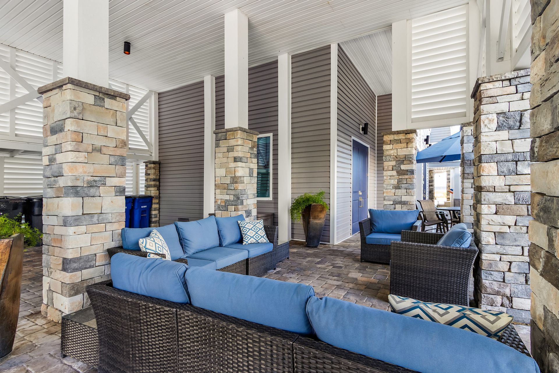 Covered outdoor patio with wicker seating, blue cushions, stone columns, and a pool view