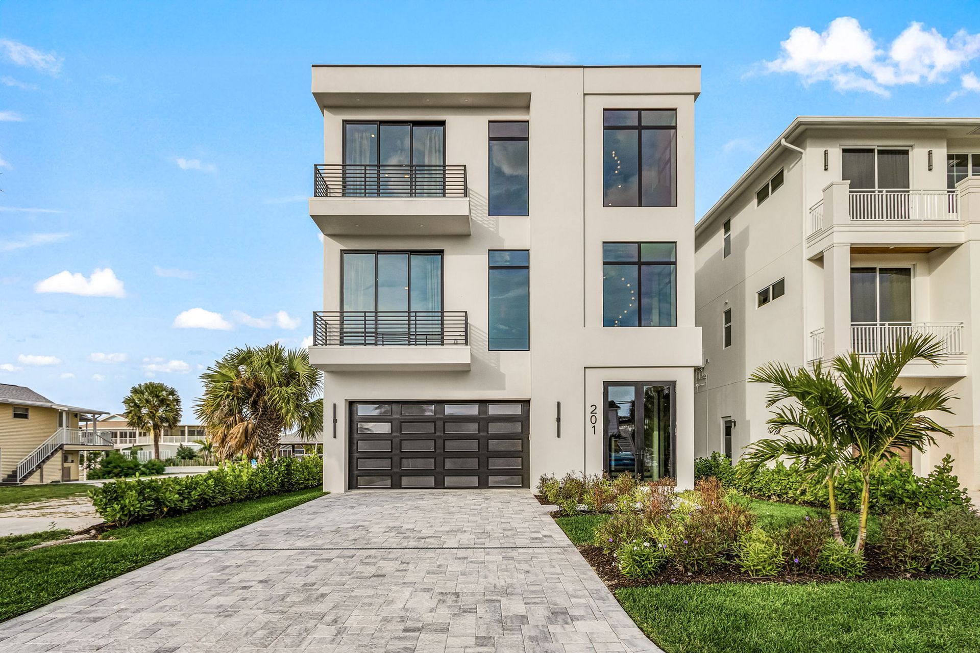 Modern three-story house with a garage and small balcony, on a paved driveway with landscaping.