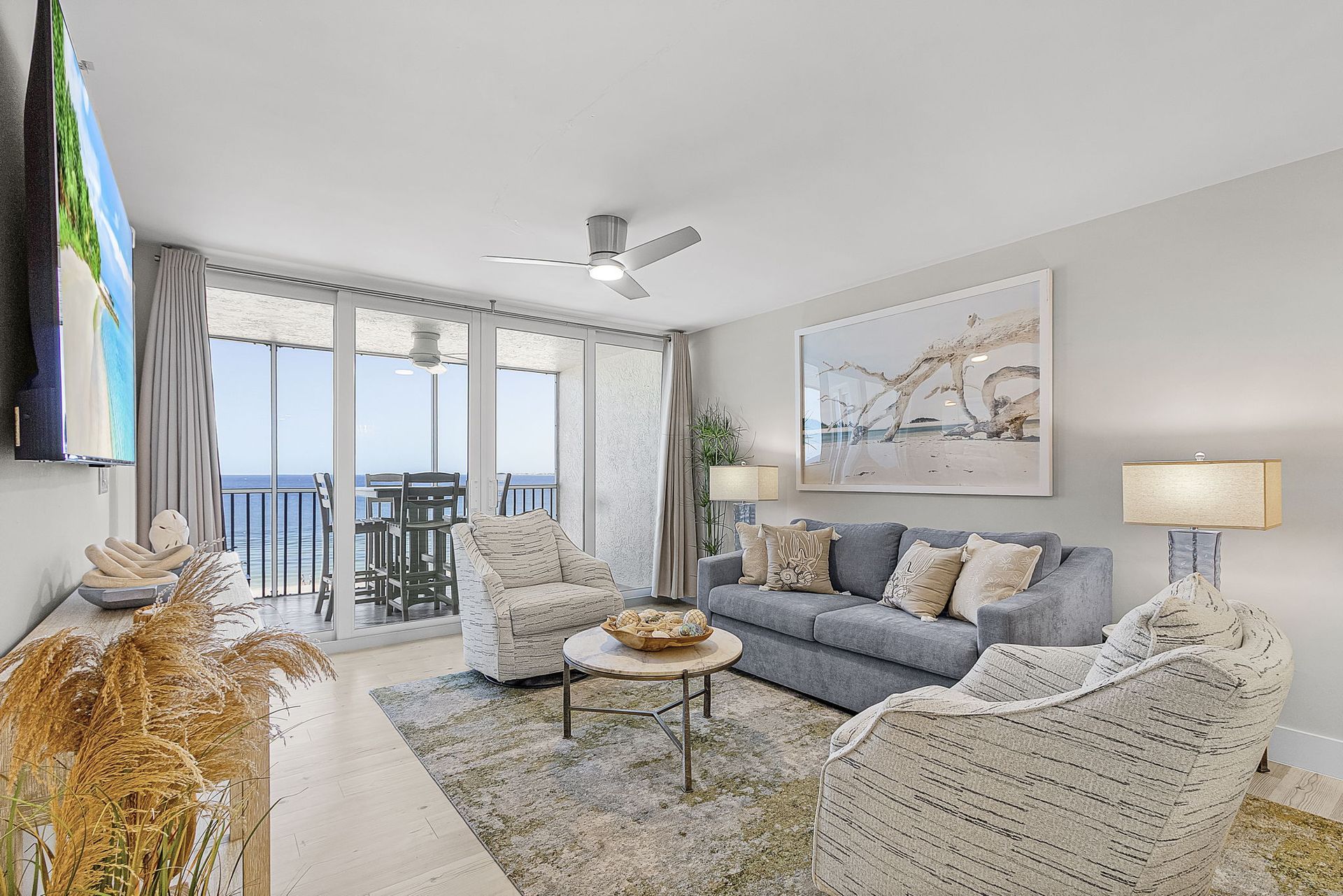Living room with ocean view, blue sofa, patterned armchairs, artwork, and ceiling fan.