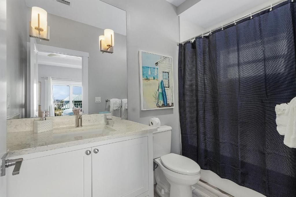 Bathroom with white vanity, toilet, blue shower curtain, and artwork; lit by sconces.