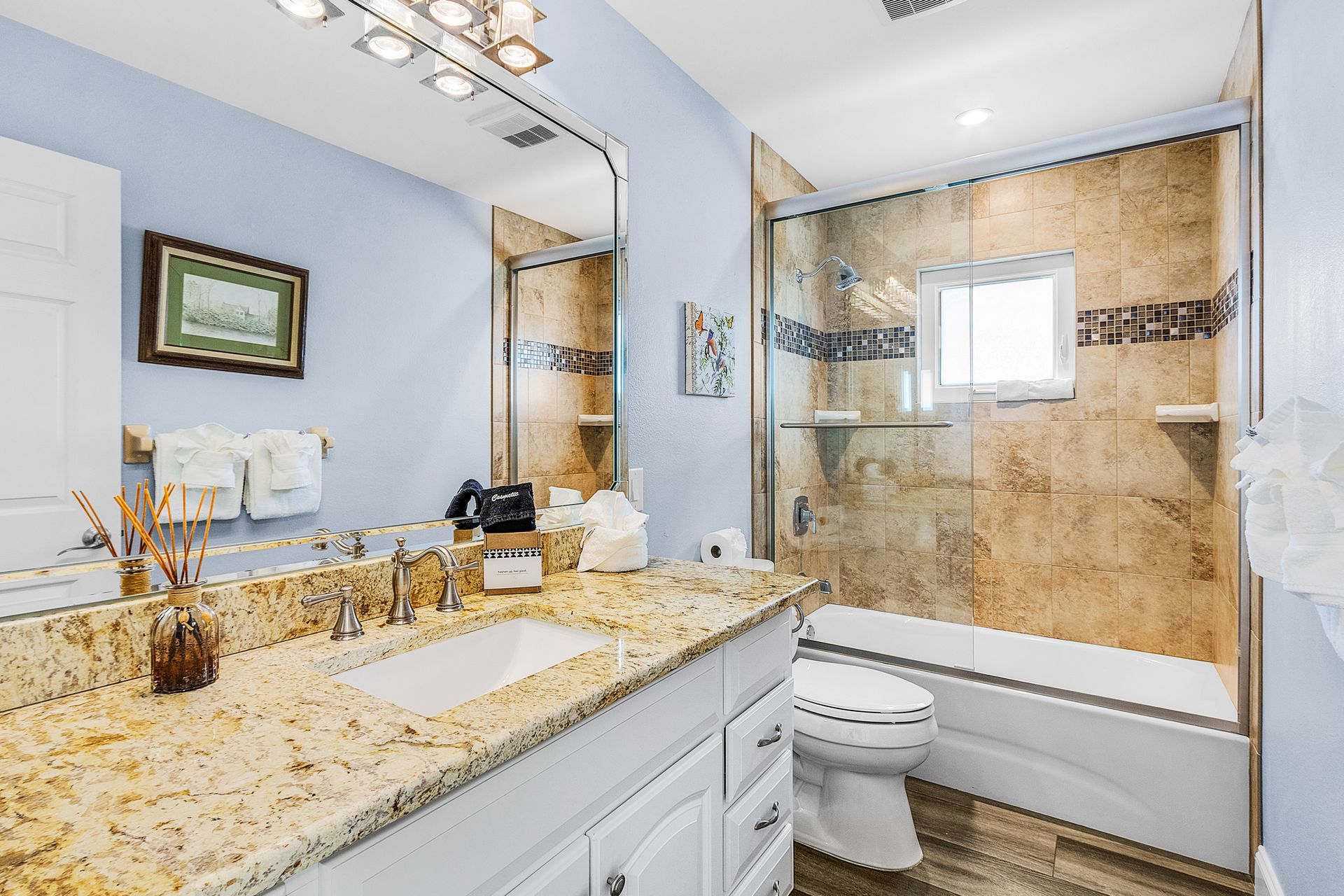 Bright bathroom with granite vanity, white cabinets, mirror, toilet, and glass-enclosed shower with beige tile.