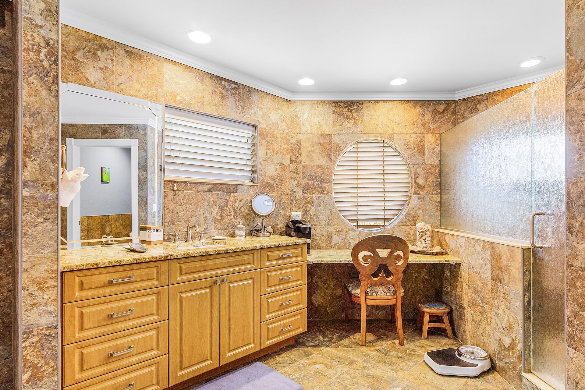 Bright bathroom with wooden vanity, round mirror, and a small wicker chair by the window.