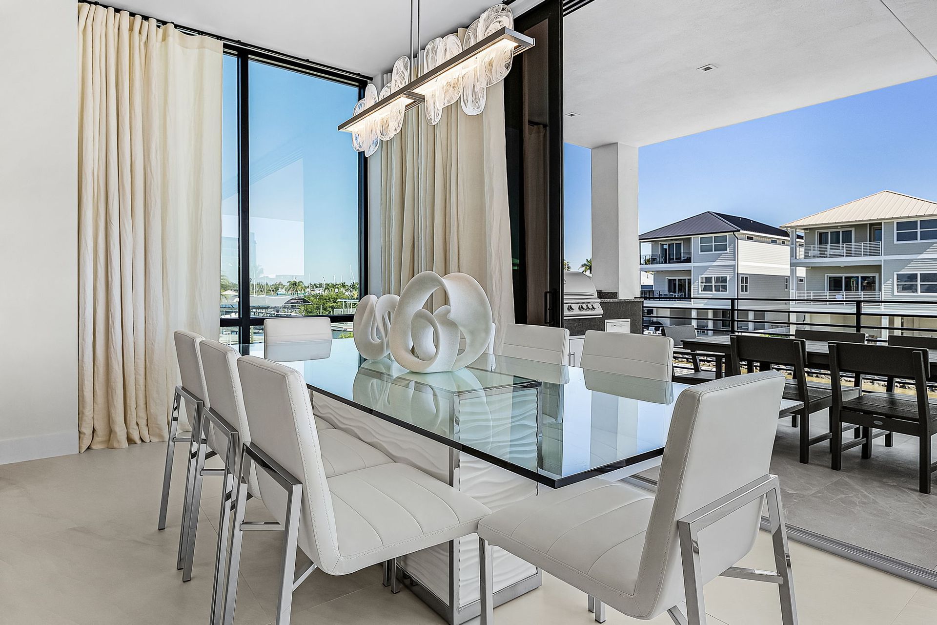 Dining room with glass table, white chairs, large window with view, and chandelier.