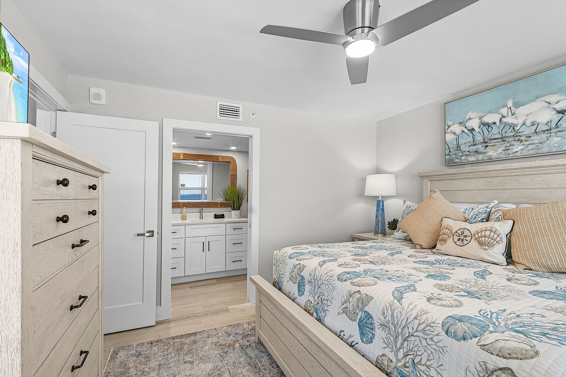 Bedroom with a white washed bed frame, floral comforter, and a chest of drawers. Bathroom entrance visible.