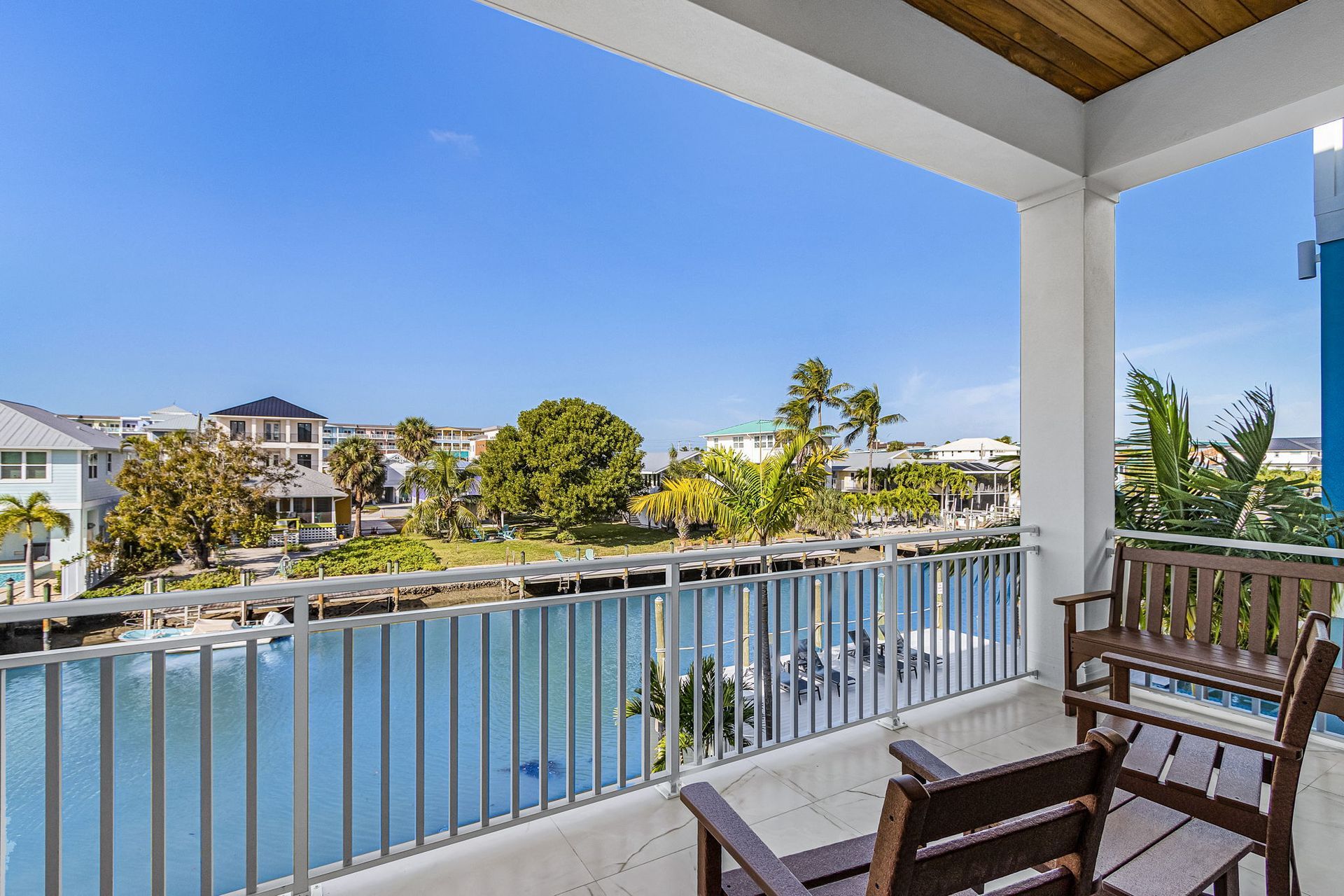 Balcony overlooking a canal with two wooden chairs. Buildings and trees visible in the background under a blue sky.