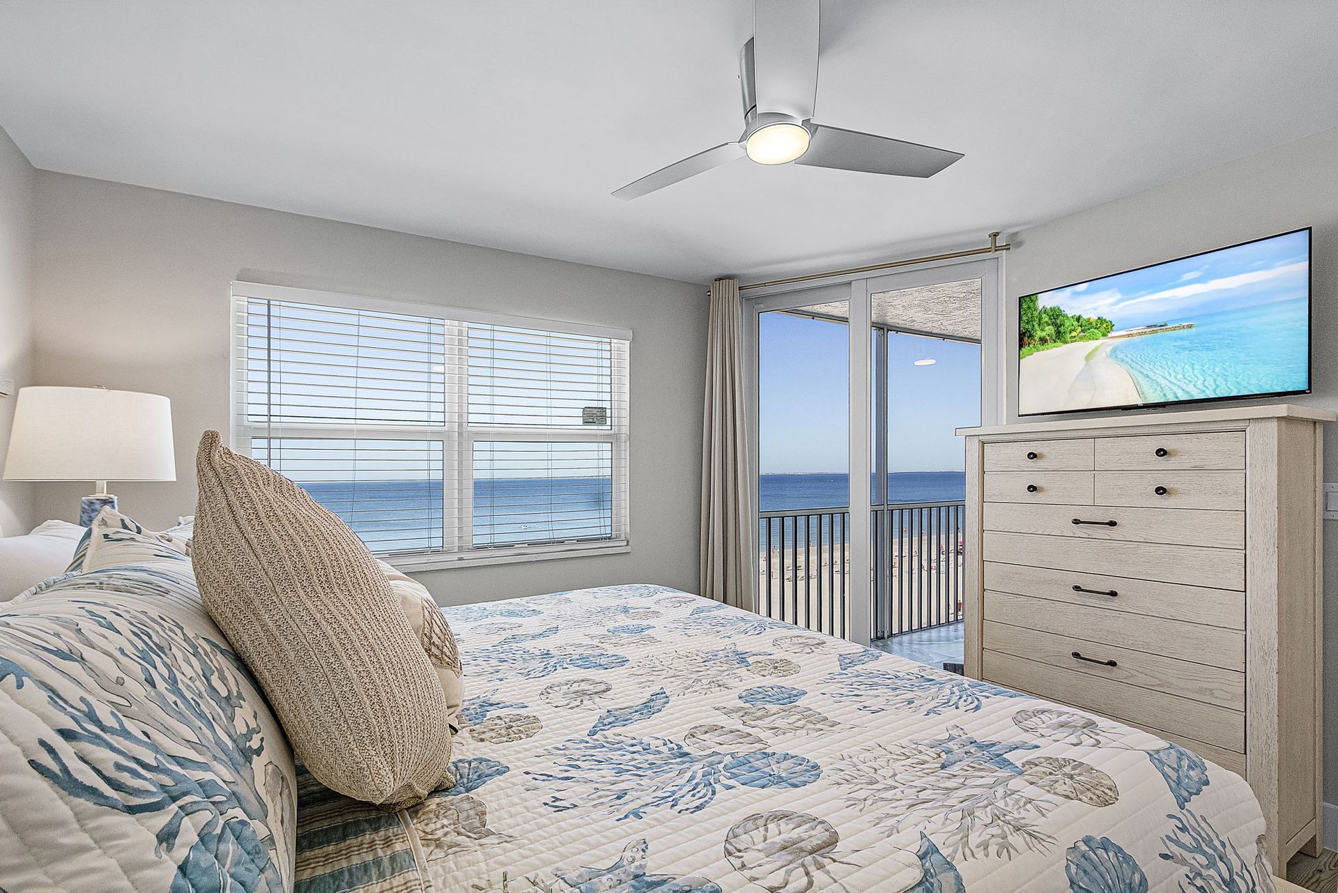 Bedroom with ocean view; bed with blue floral quilt; large window and balcony with water view; TV.