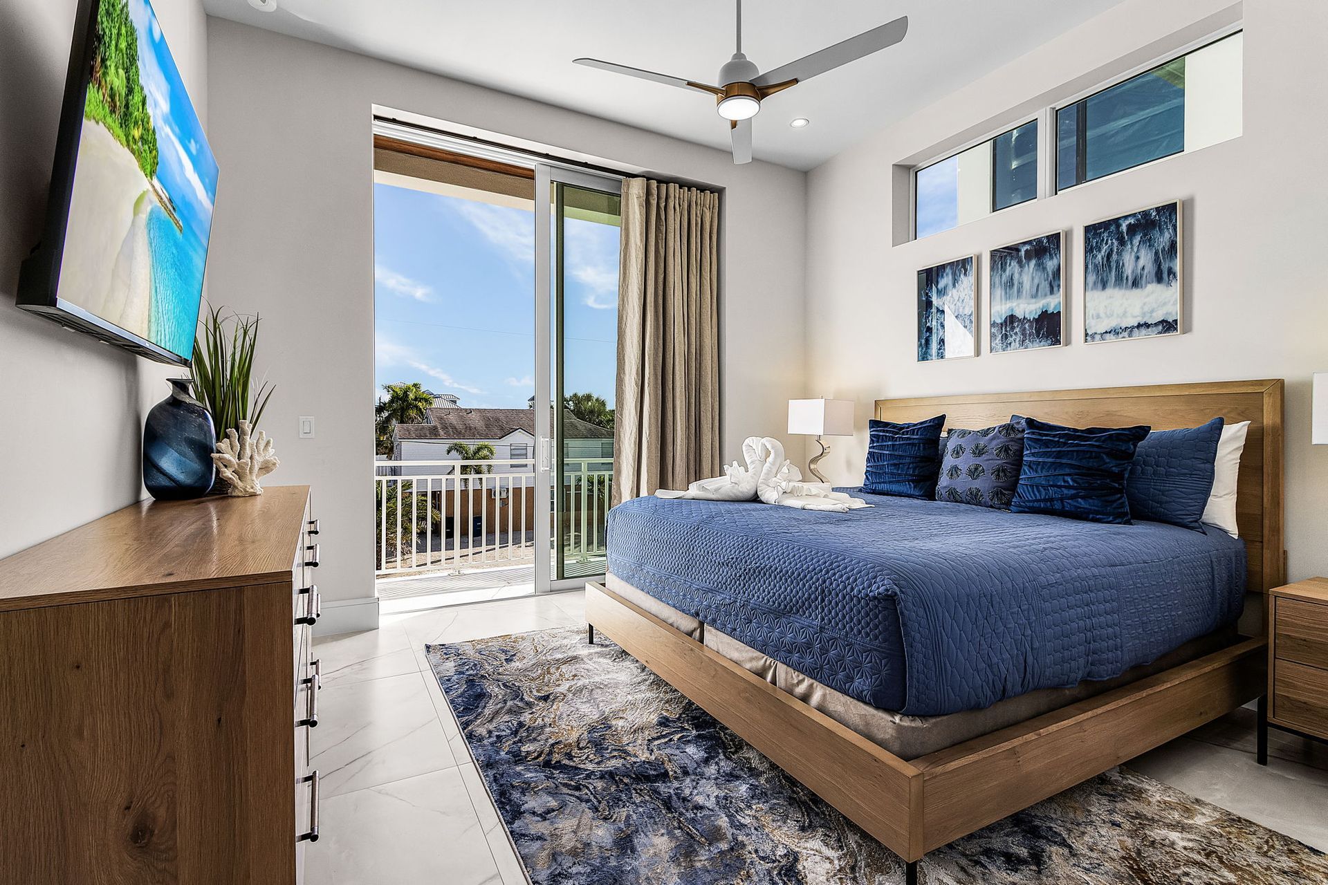 Bedroom with king-size bed, balcony access, TV, and artwork. Blue and neutral tones. Bright, airy setting.