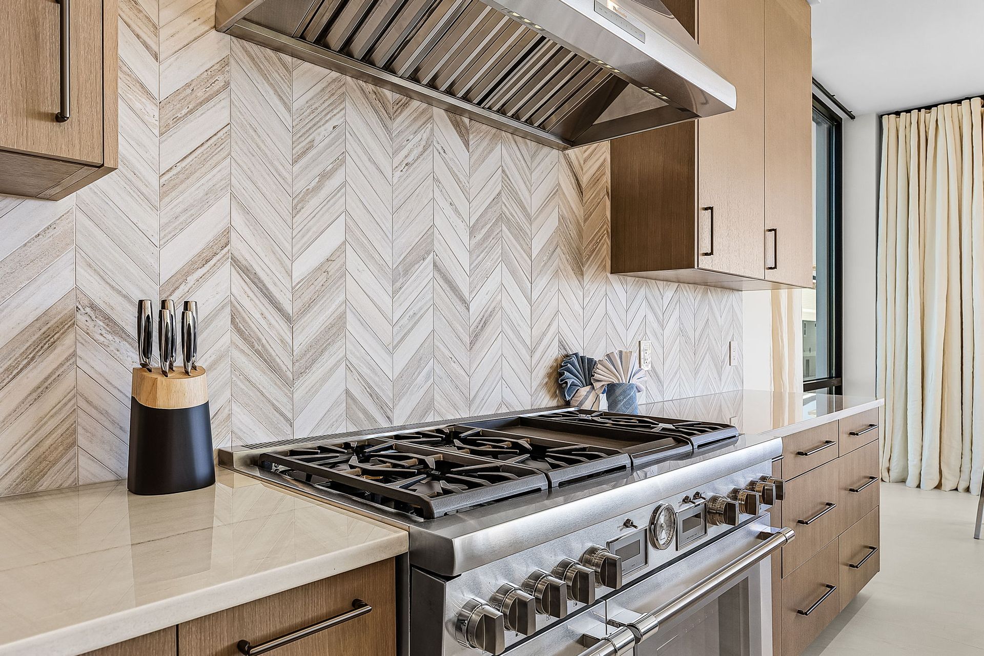 Modern kitchen with stainless steel range, light wood cabinets, and herringbone tile backsplash.