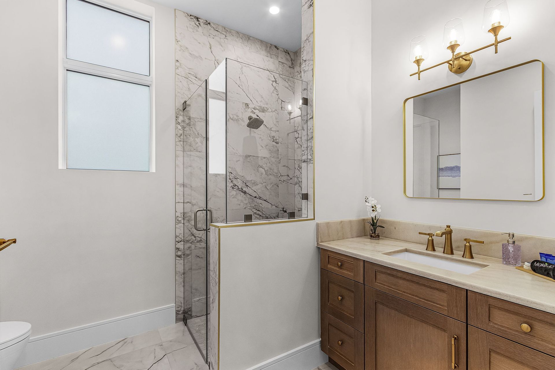 Bathroom with marble shower, wood vanity, gold fixtures, and a window.