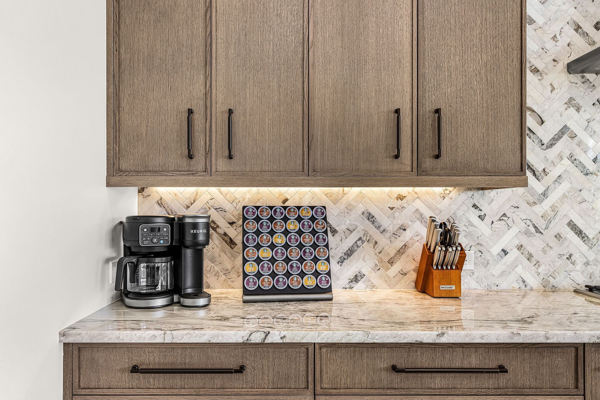 Kitchen cabinets, countertop, and backsplash in a warm-toned, modern style. Appliances and a patterned towel are visible.