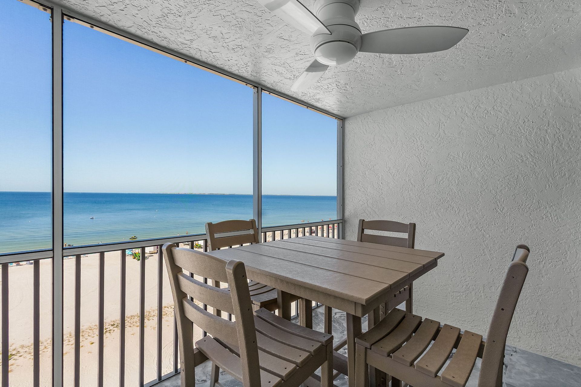 A screened-in porch with a table and chairs overlooking a beach and ocean. Blue sky, white sand.