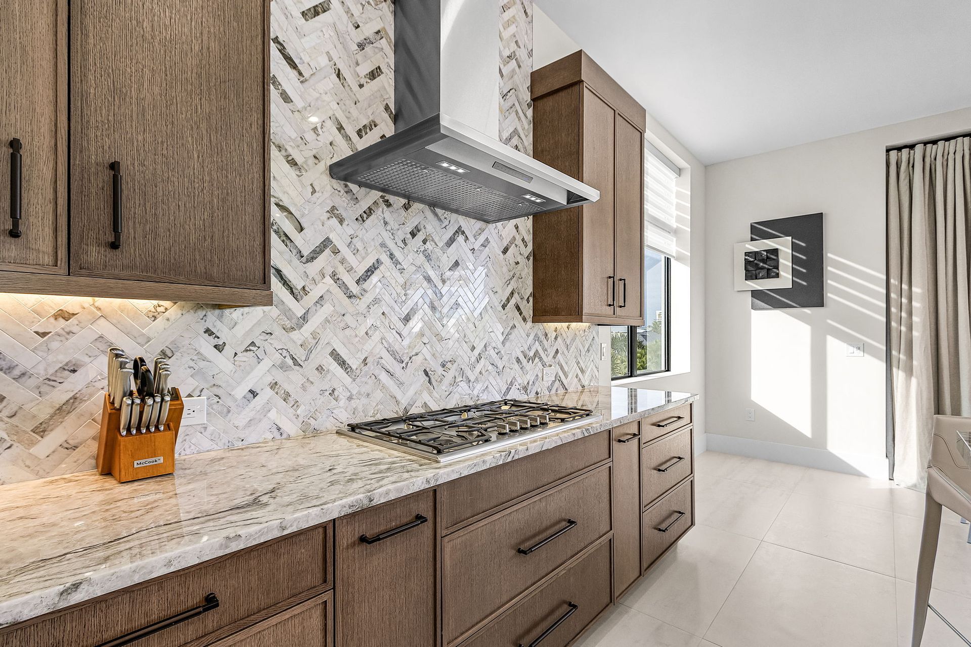 Kitchen with brown cabinets, white and gray backsplash, stainless steel range hood, and white countertops.