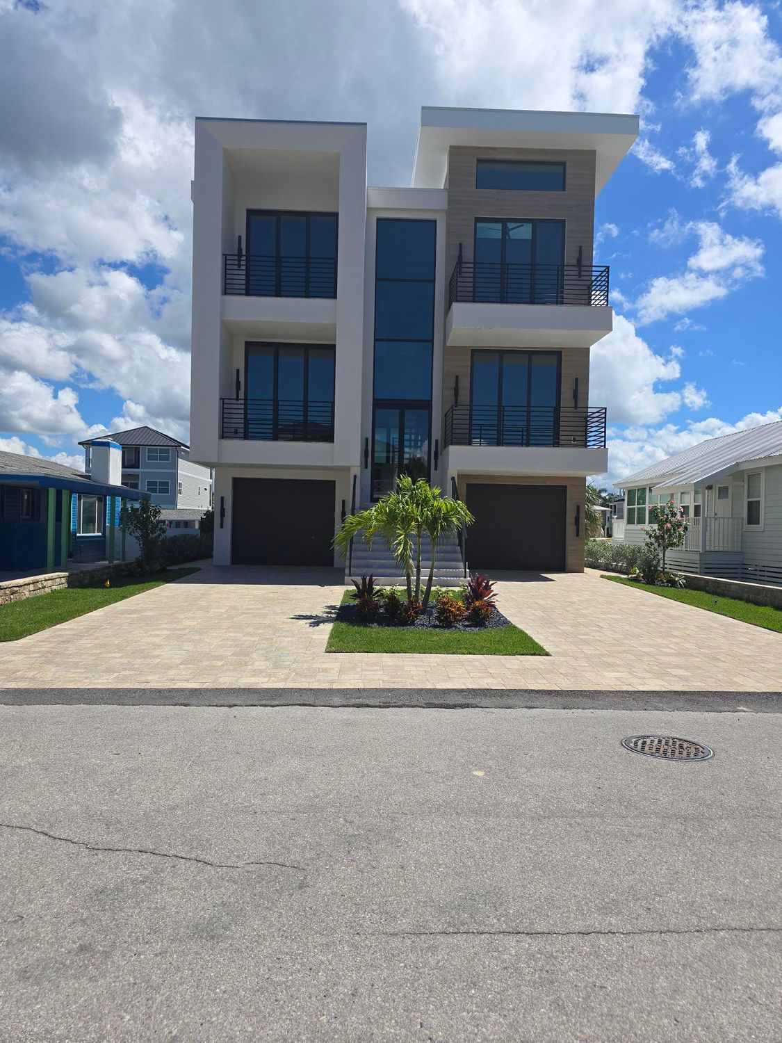 Modern multi-story house with tan facade, large windows, and two garage doors. Palm tree in front.