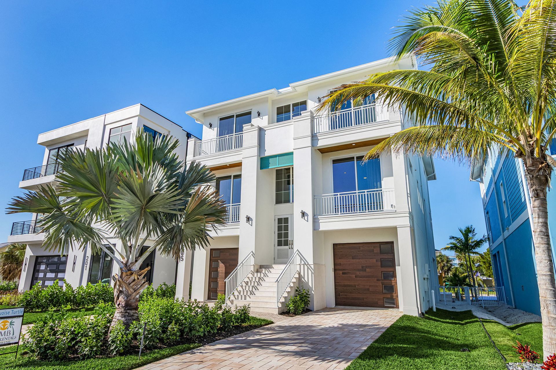 White multi-story house with palm trees, blue sky, and a stone driveway.