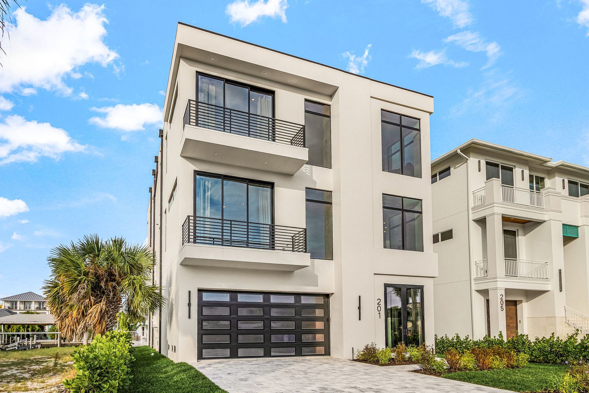 Modern three-story house with black-framed windows, balconies, and a garage door. Blue sky with clouds.