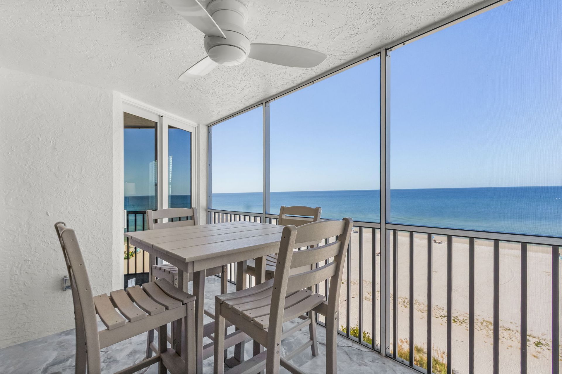 Balcony with table and chairs overlooking a beach and ocean. Blue sky, white walls, and a ceiling fan.