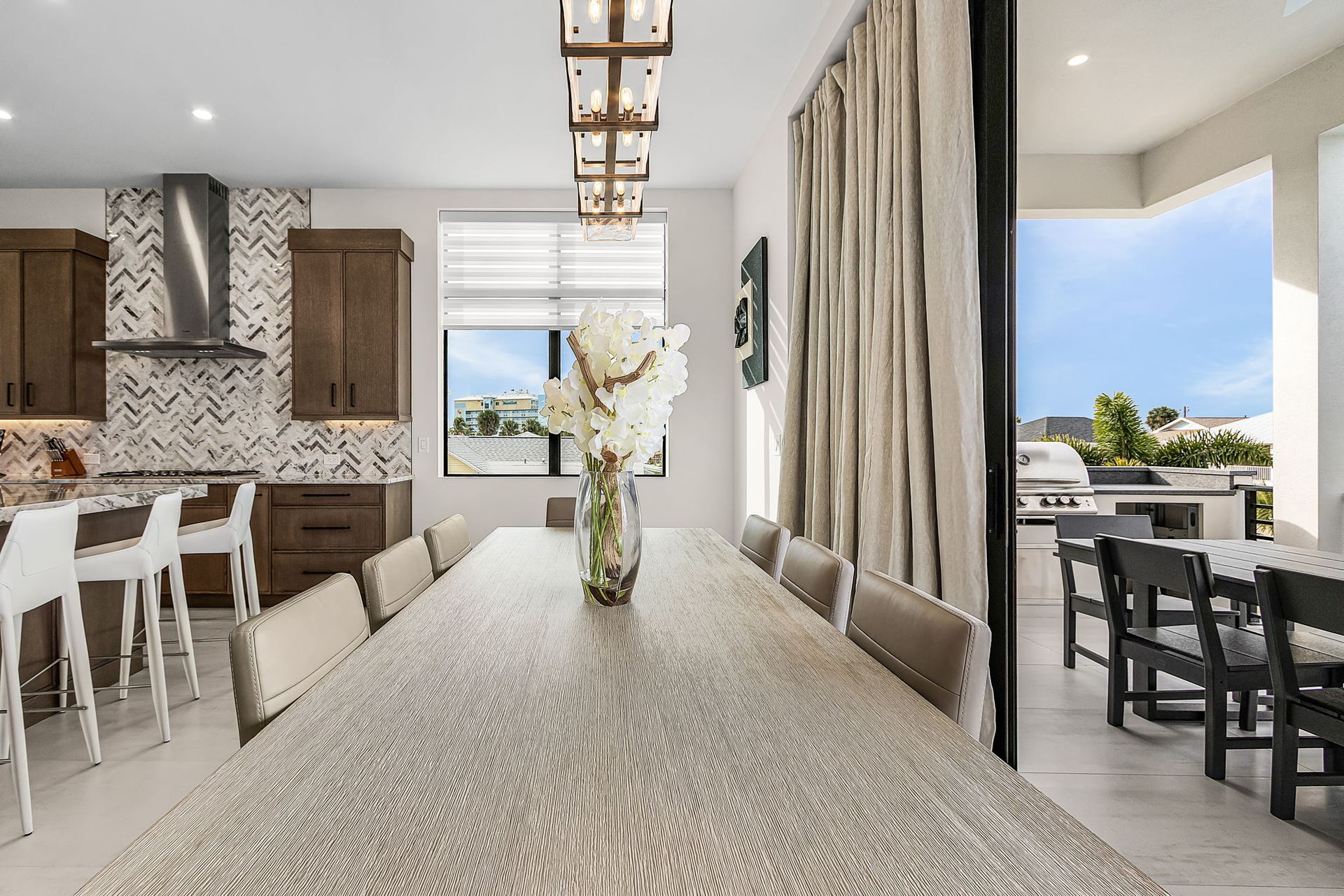 A dining room with a long table set with chairs, a kitchen, and a patio with an outdoor grill.