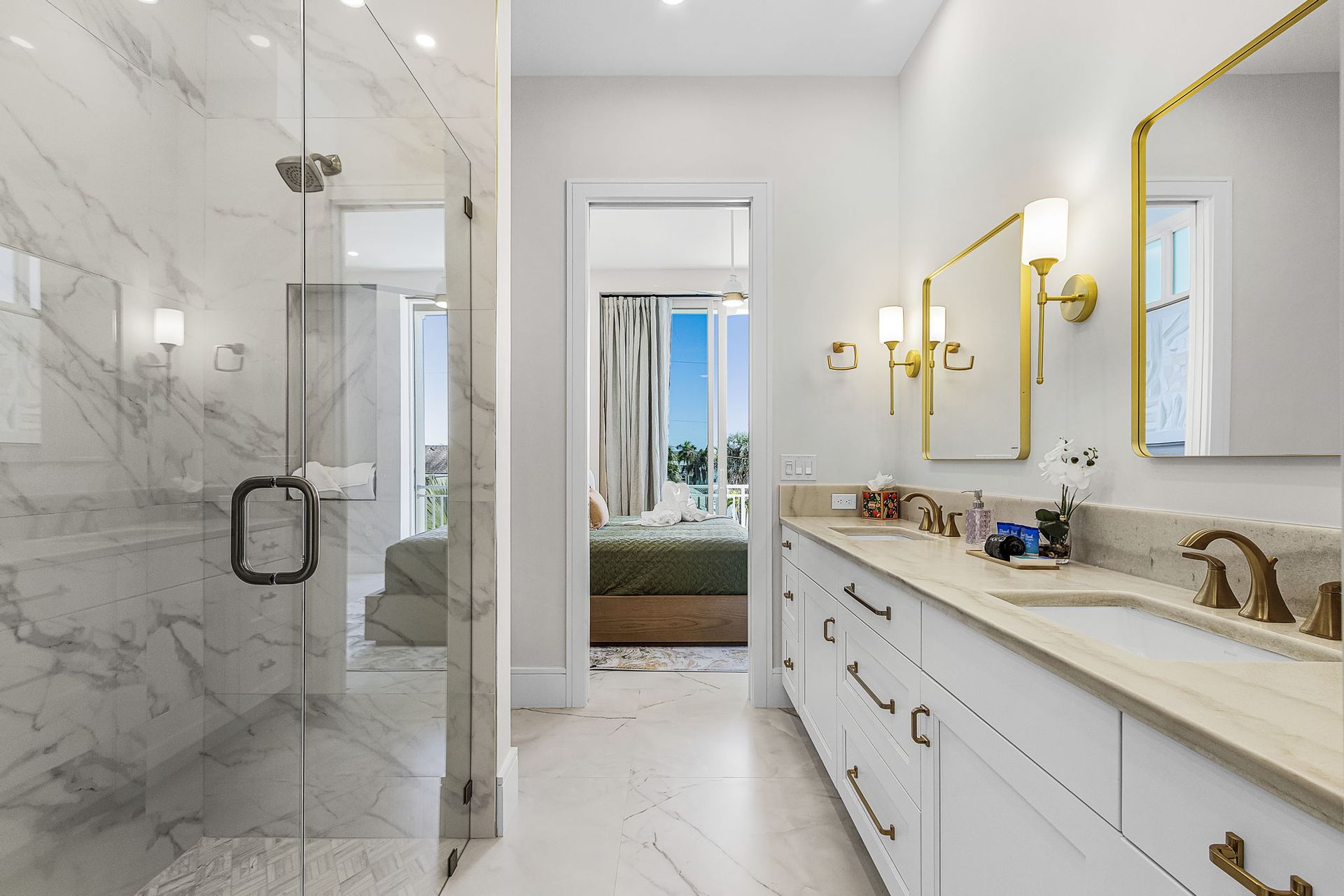 Luxurious white marble bathroom with a glass shower and gold accents, leading to a bedroom.