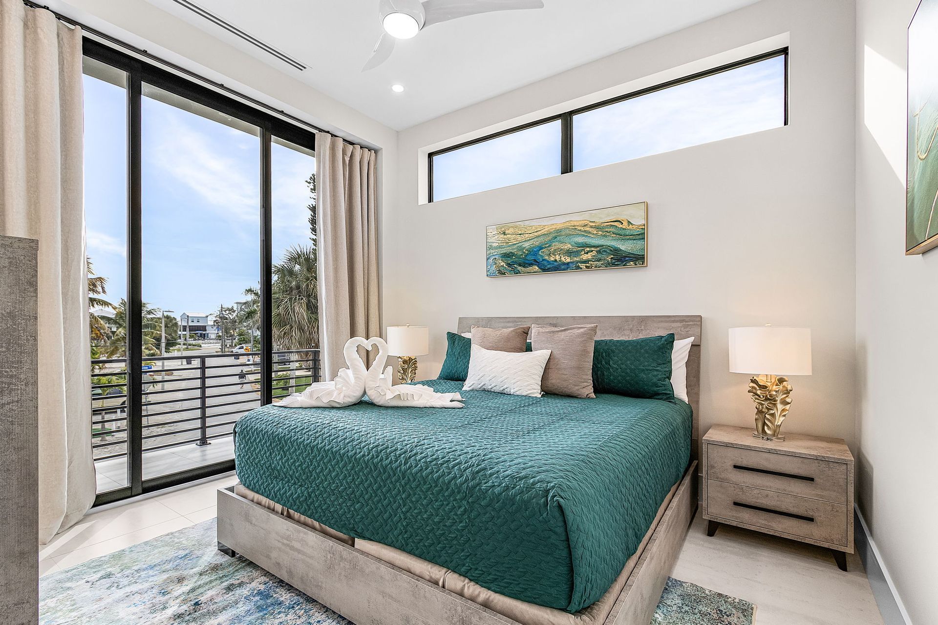 Bedroom with teal bedspread, large windows, and balcony overlooking a sunny outdoor view.