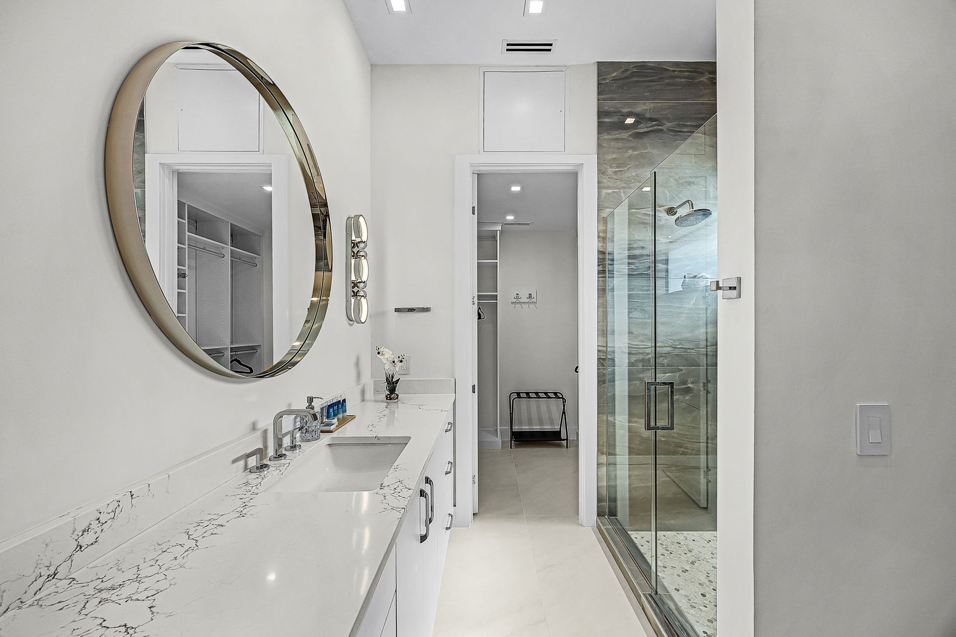 White bathroom with marble countertop, round mirror, and glass shower.
