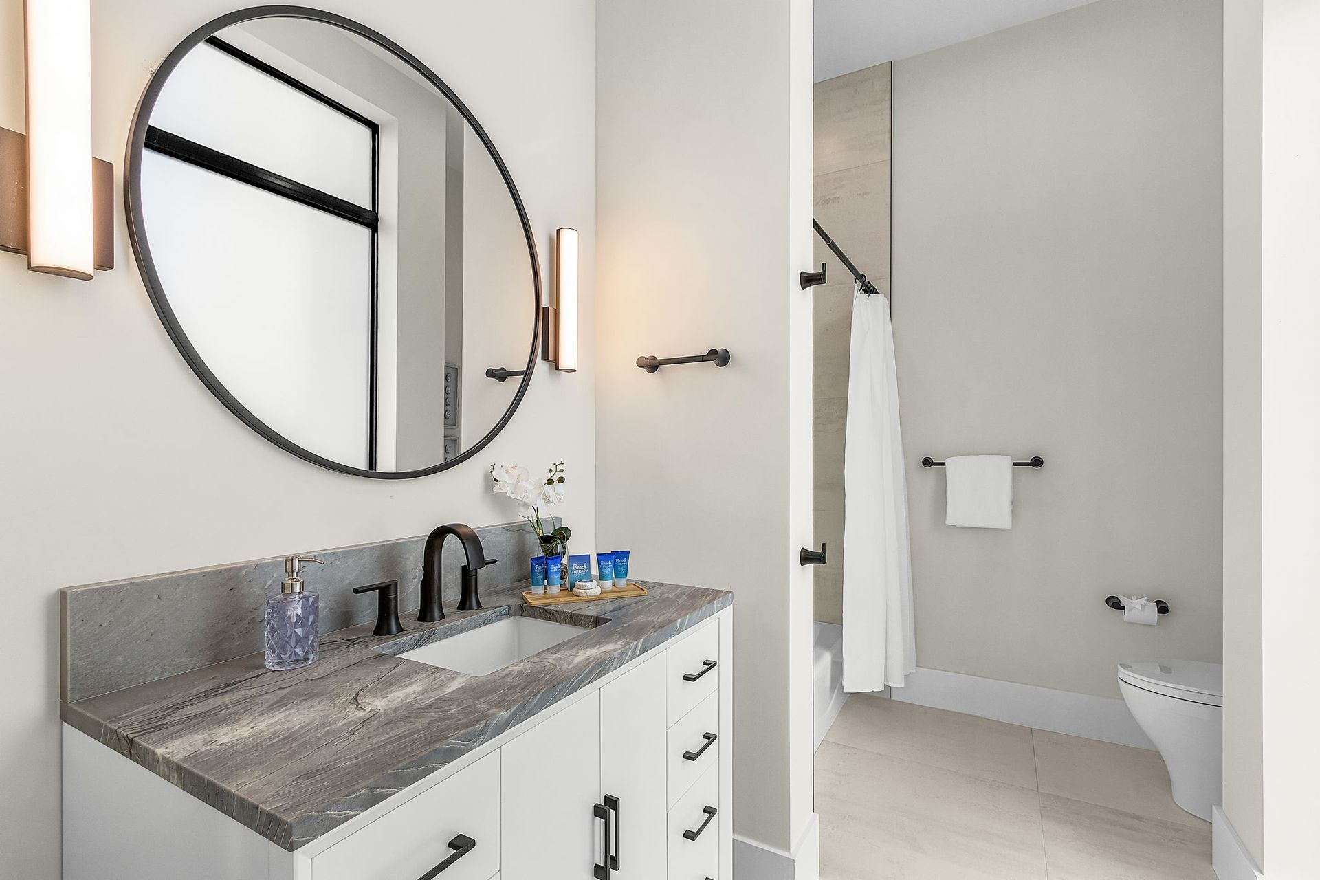 Bathroom with round mirror, white vanity, gray countertop, and open doorway to shower.