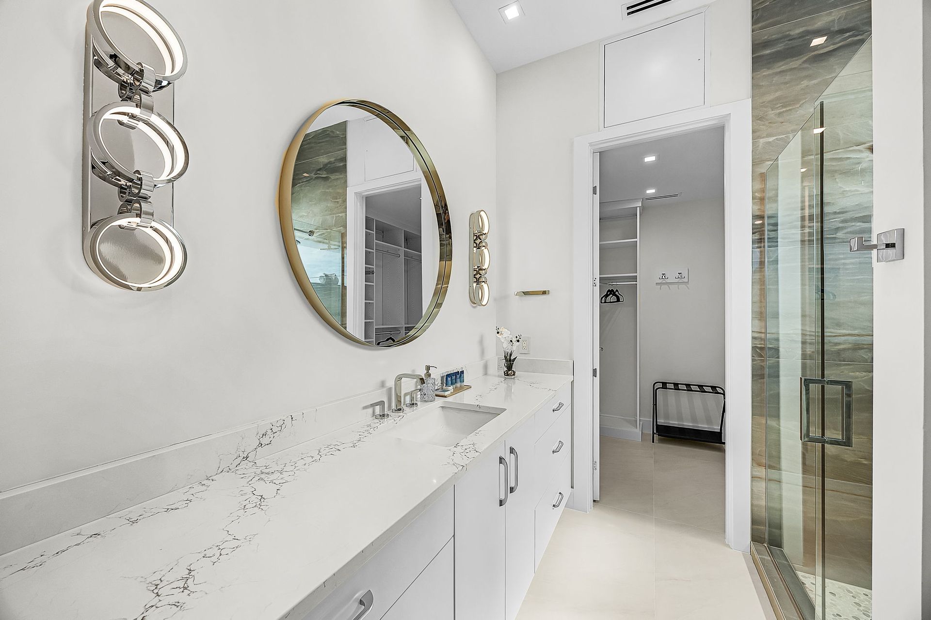 Bright white bathroom with marble counter, oval mirror, and three-light sconce. Walk-in closet visible.