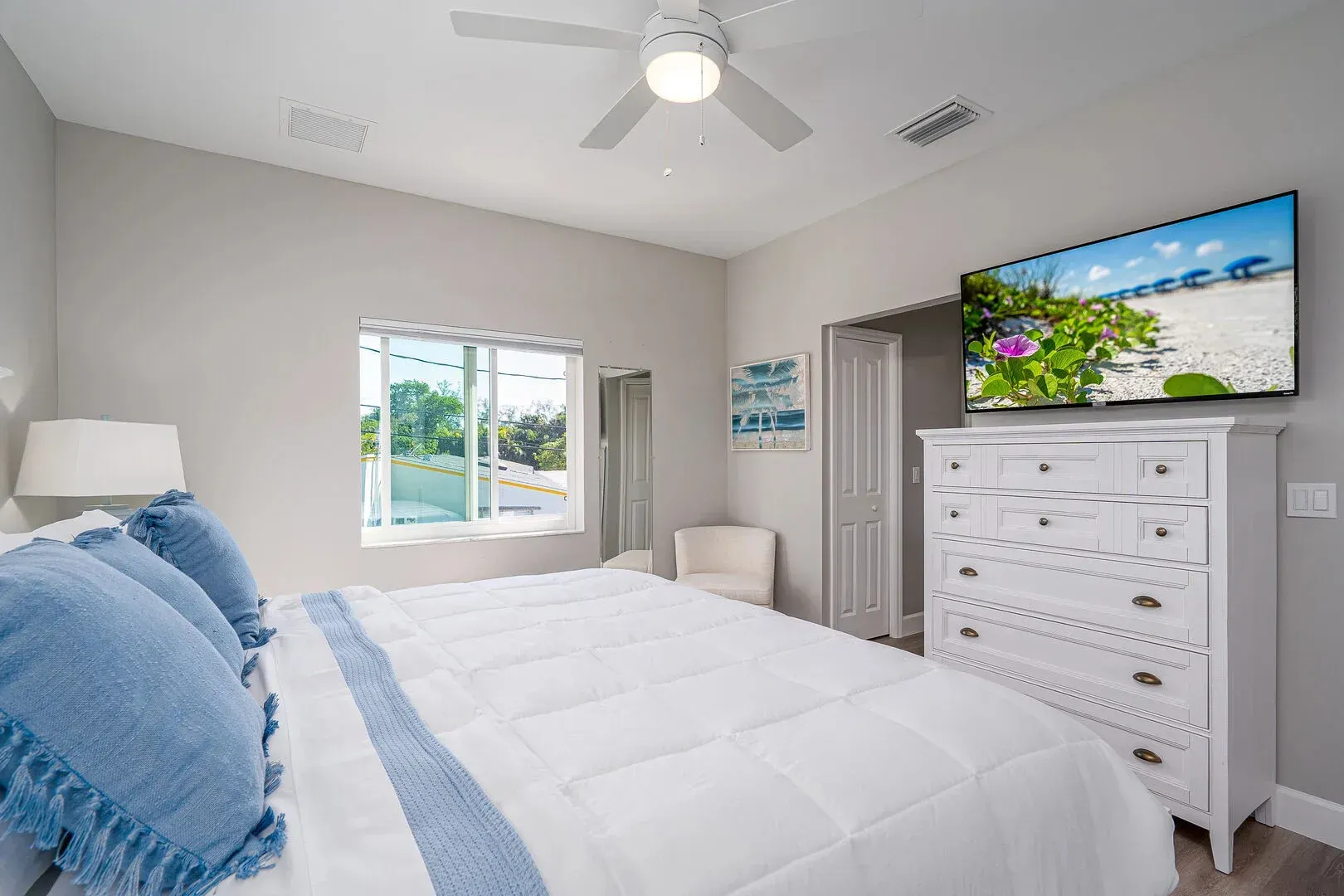 Bedroom with a white bed, TV, dresser, and a window. Light blue accents and neutral walls.