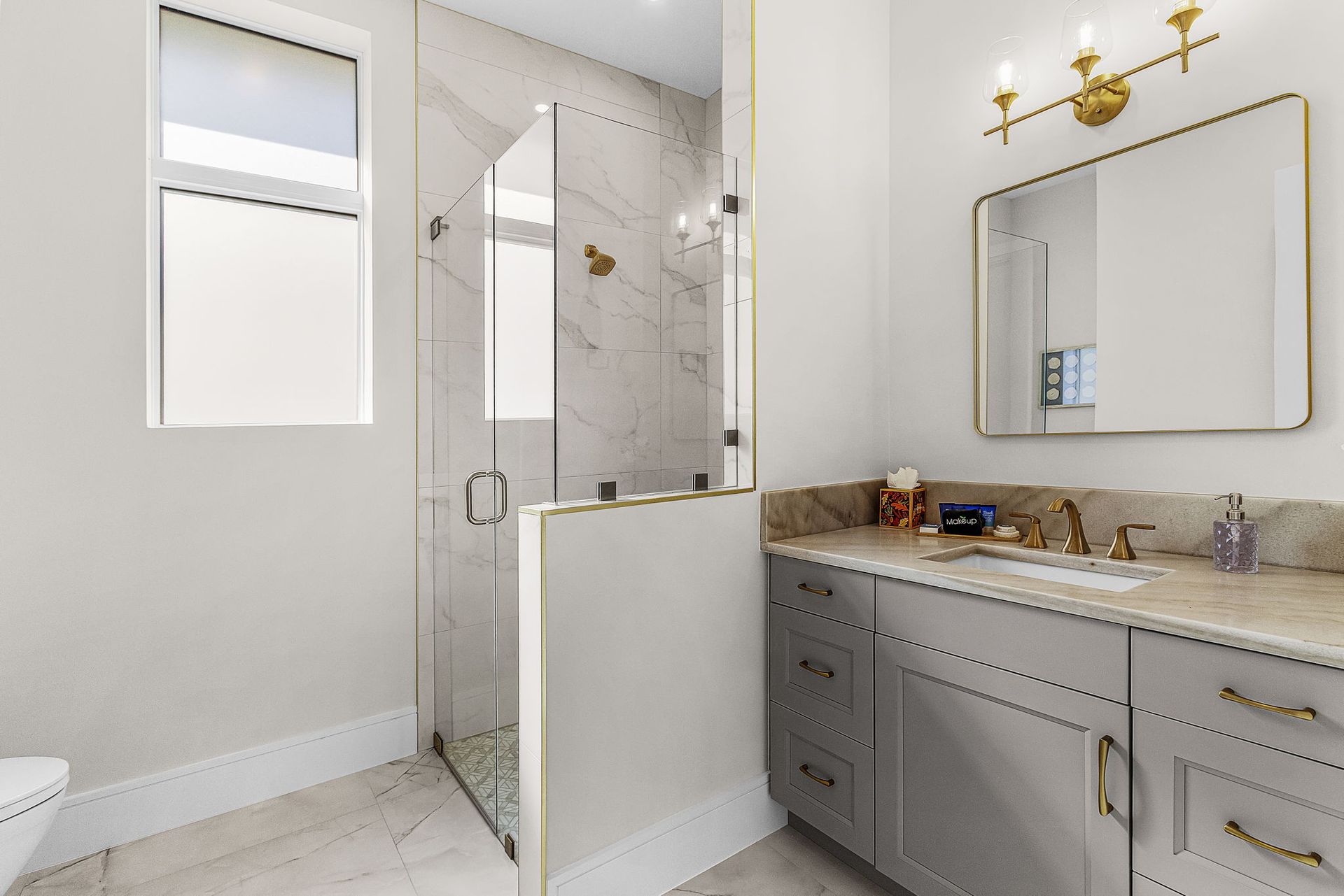 Bathroom with gray vanity, gold fixtures, marble shower, and a window.