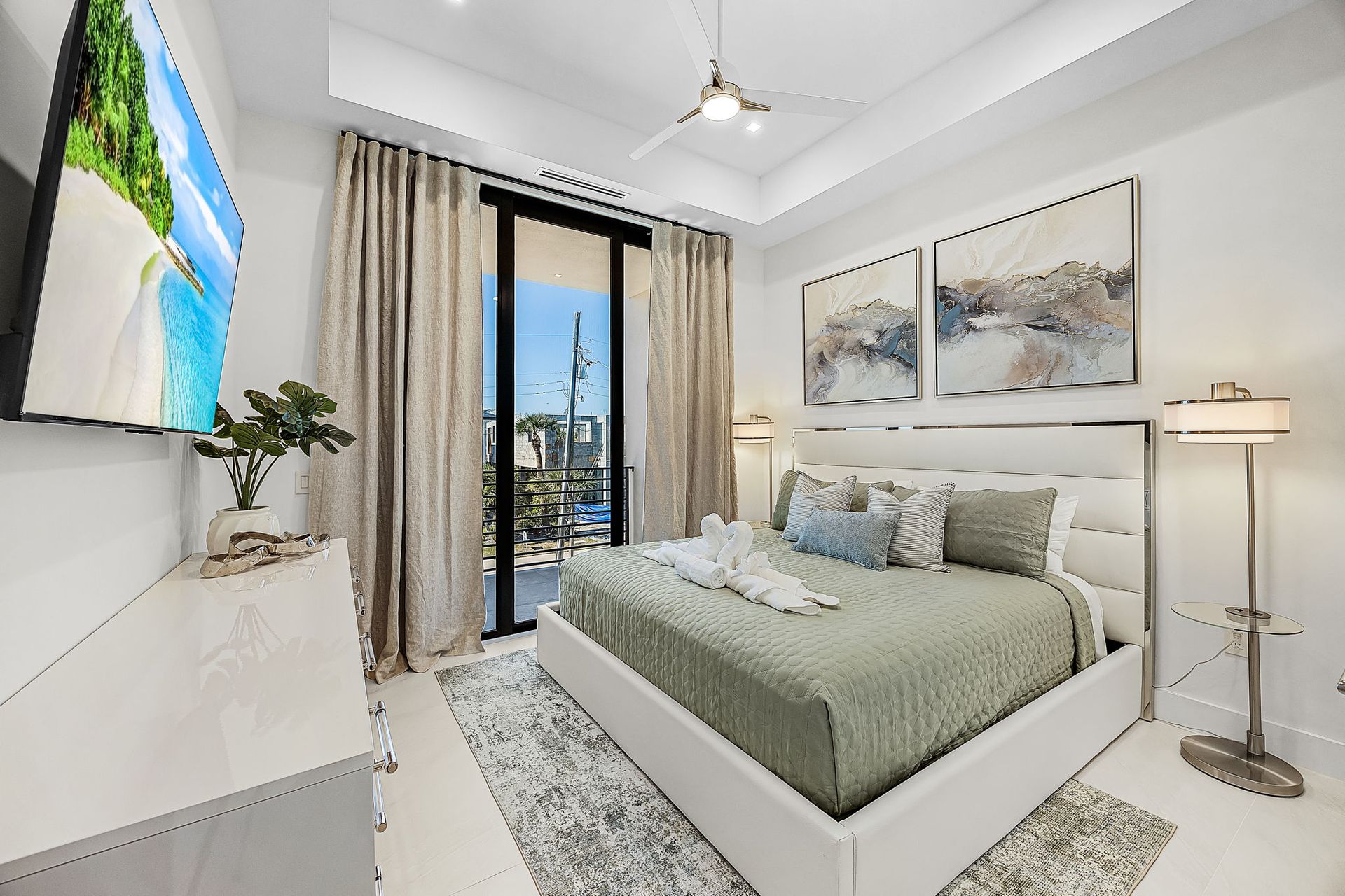 Bright bedroom with white walls, bed, and dresser. Large window with sheer curtains, artwork, and TV.