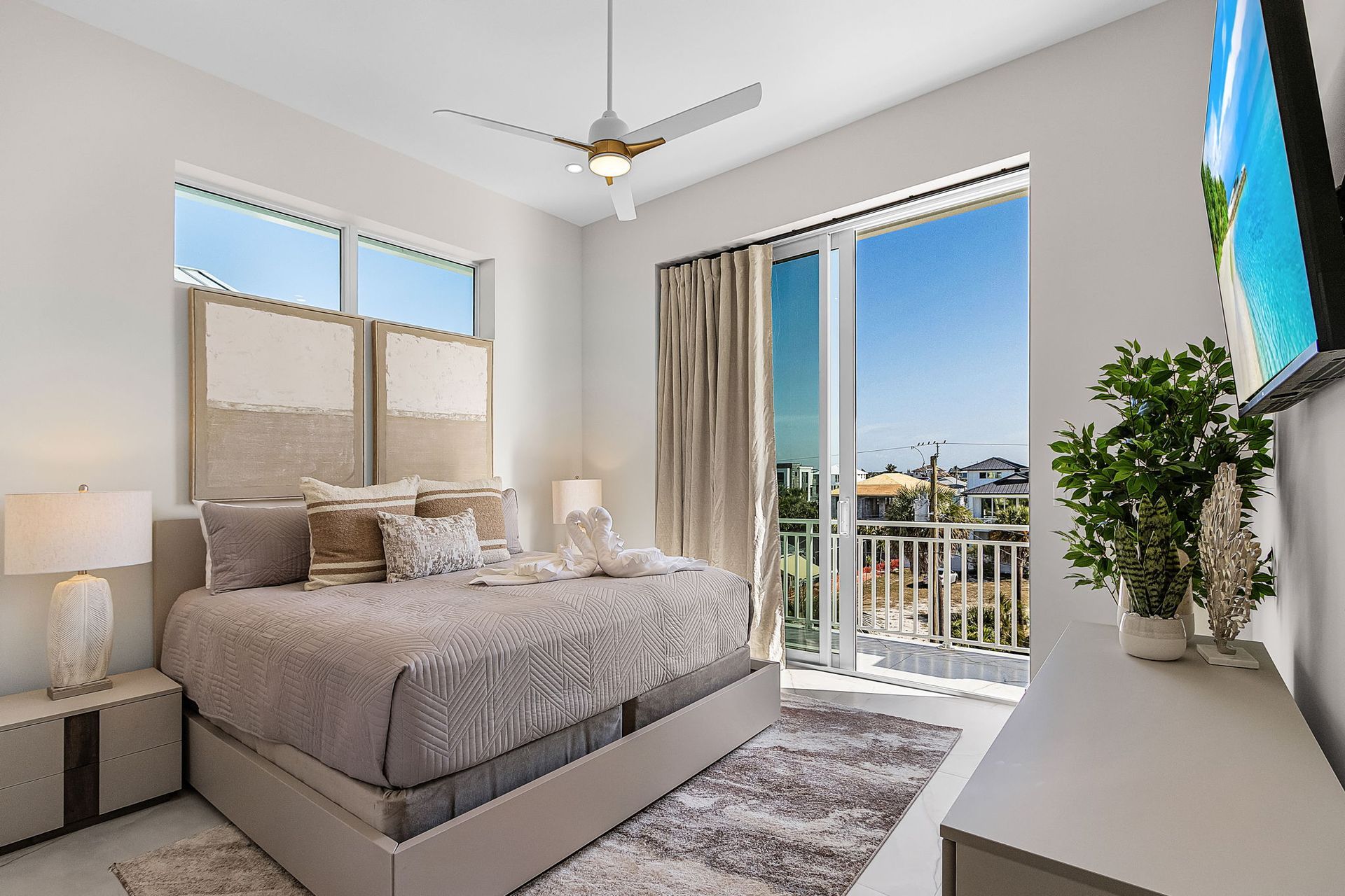 Bedroom with bed, balcony view, neutral tones, sliding glass door, TV on dresser, and decorative plants.
