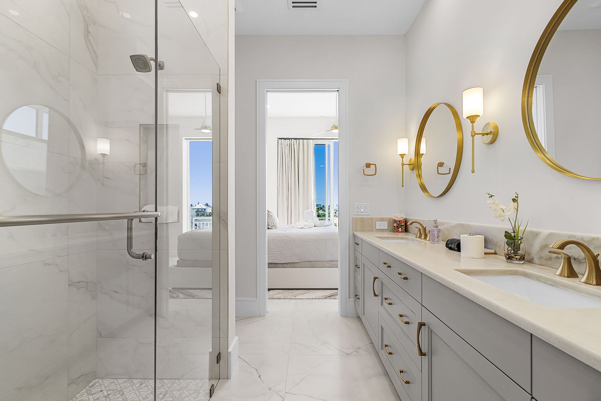 Bright, modern bathroom with glass shower, double vanity, and doorway leading to a bedroom.