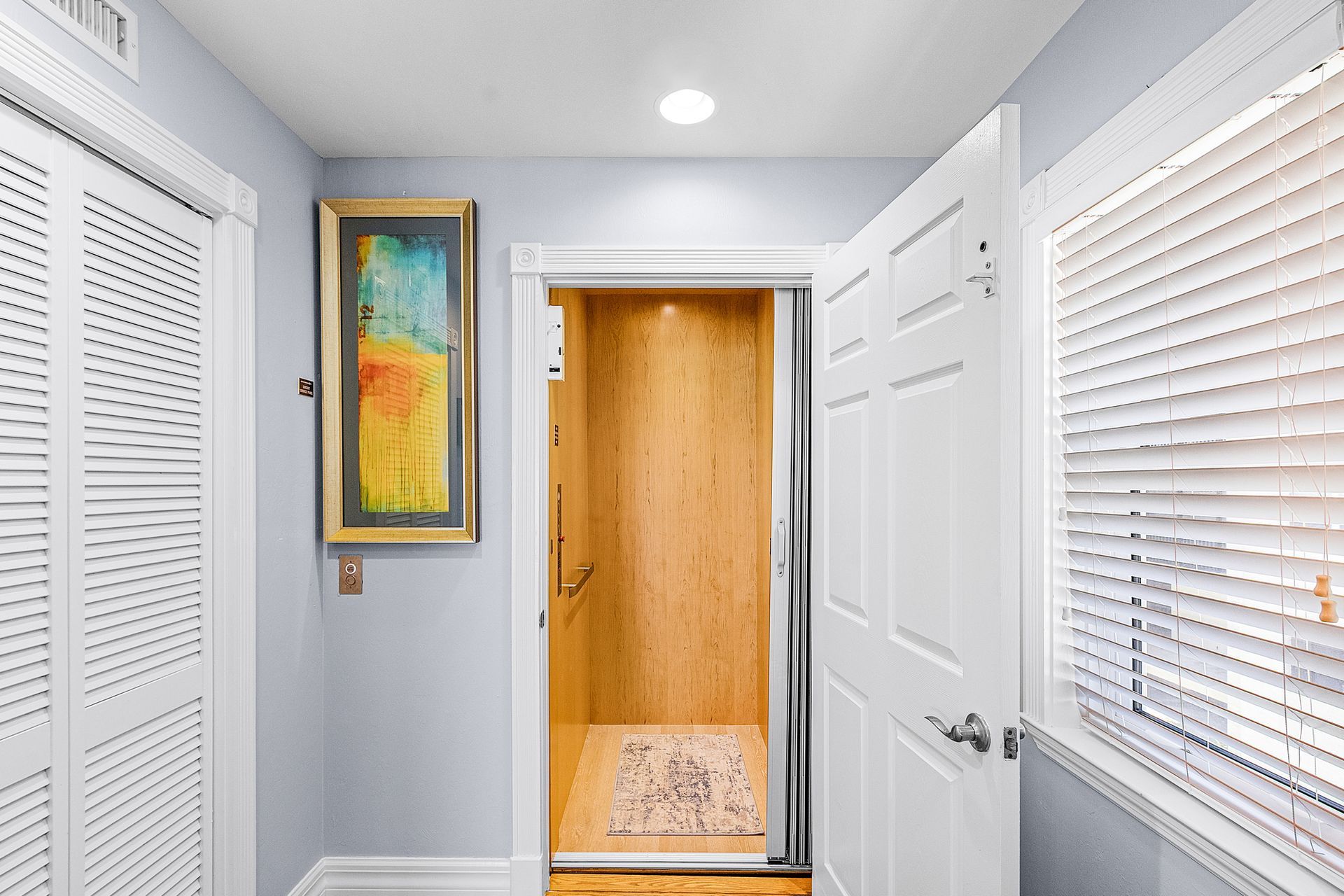 Bright hallway with white doors, blinds, colorful wall art, and an open doorway to a lit room