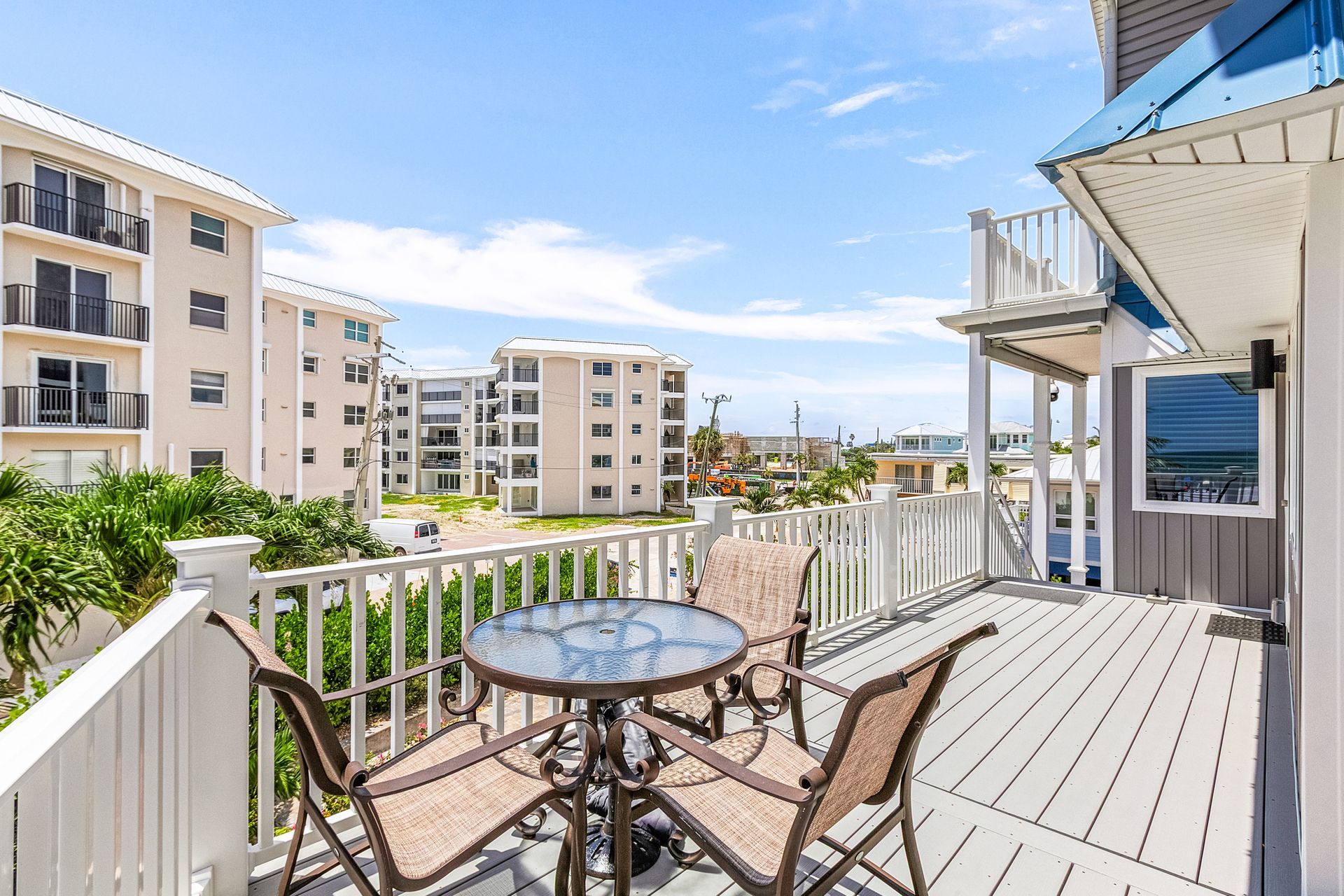 Sunny balcony with white railing, glass table, and chairs overlooking apartment buildings and blue sky