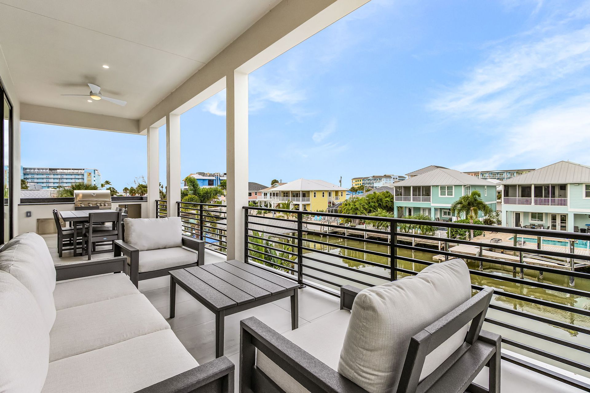Balcony with seating, table, grill, and view of waterfront homes under a blue sky.