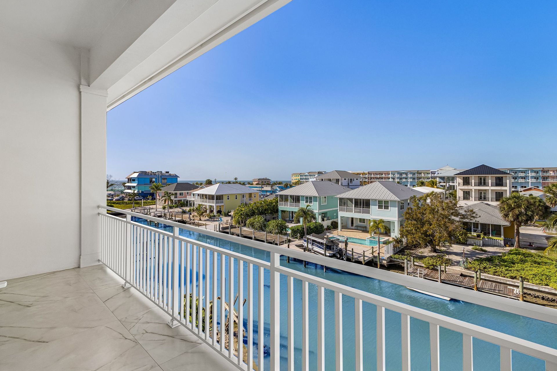 Balcony overlooking a canal with houses on a sunny day. White railing, blue water, and clear sky.