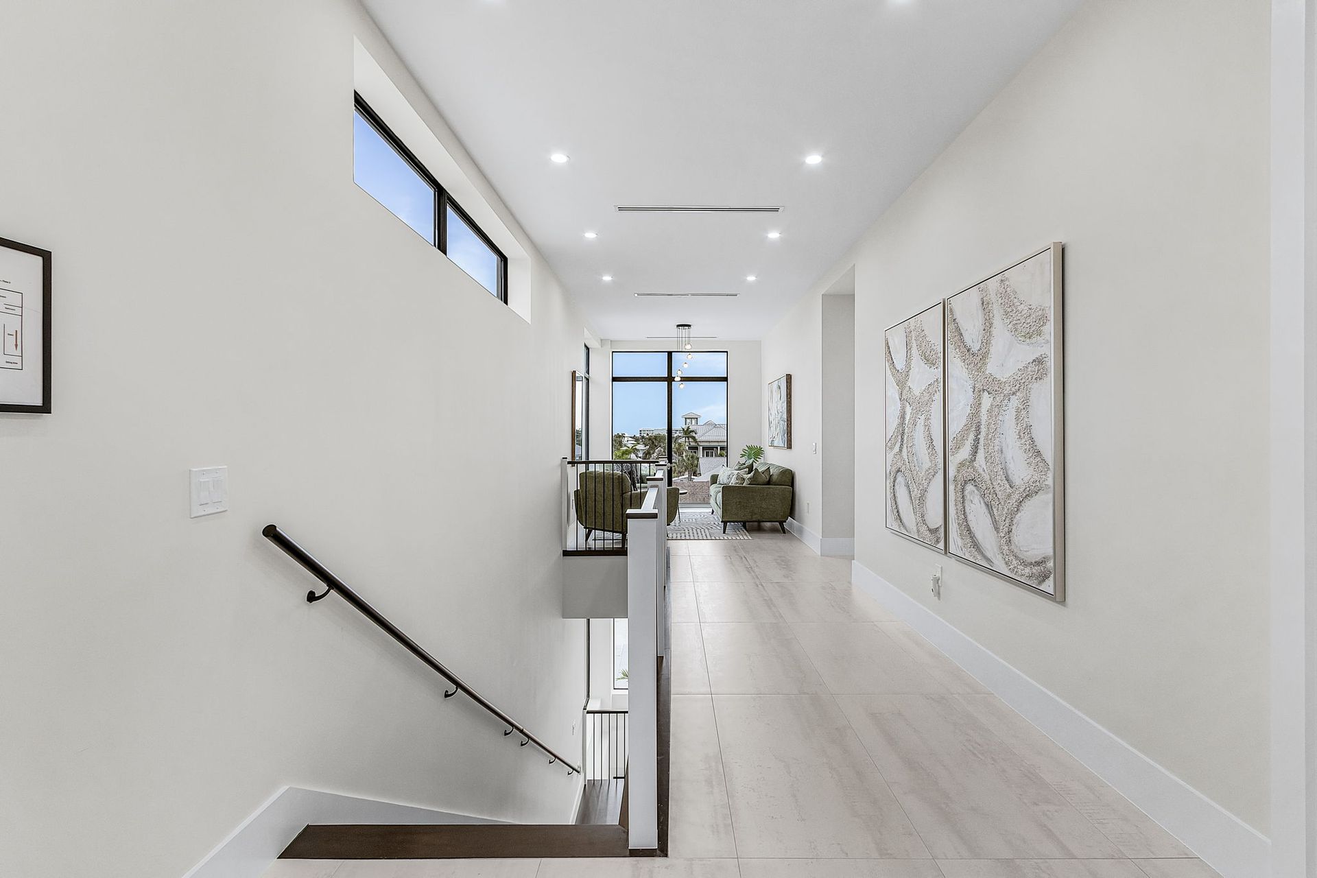 Hallway with wood floors, white walls, and stairs leading down. Large art pieces and windows.