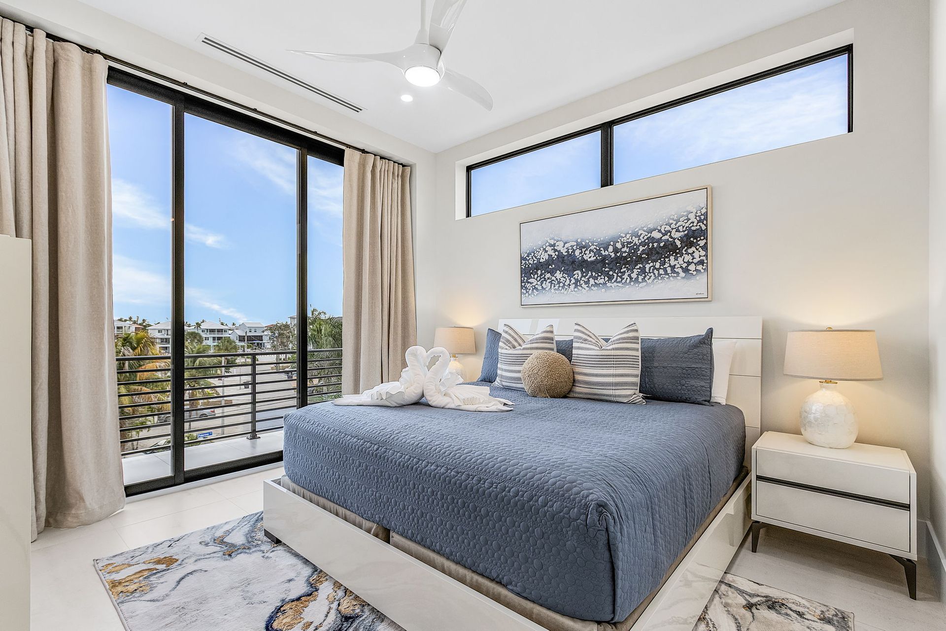 Bedroom with balcony and large windows. Blue bed, neutral walls, art, and light-colored curtains.