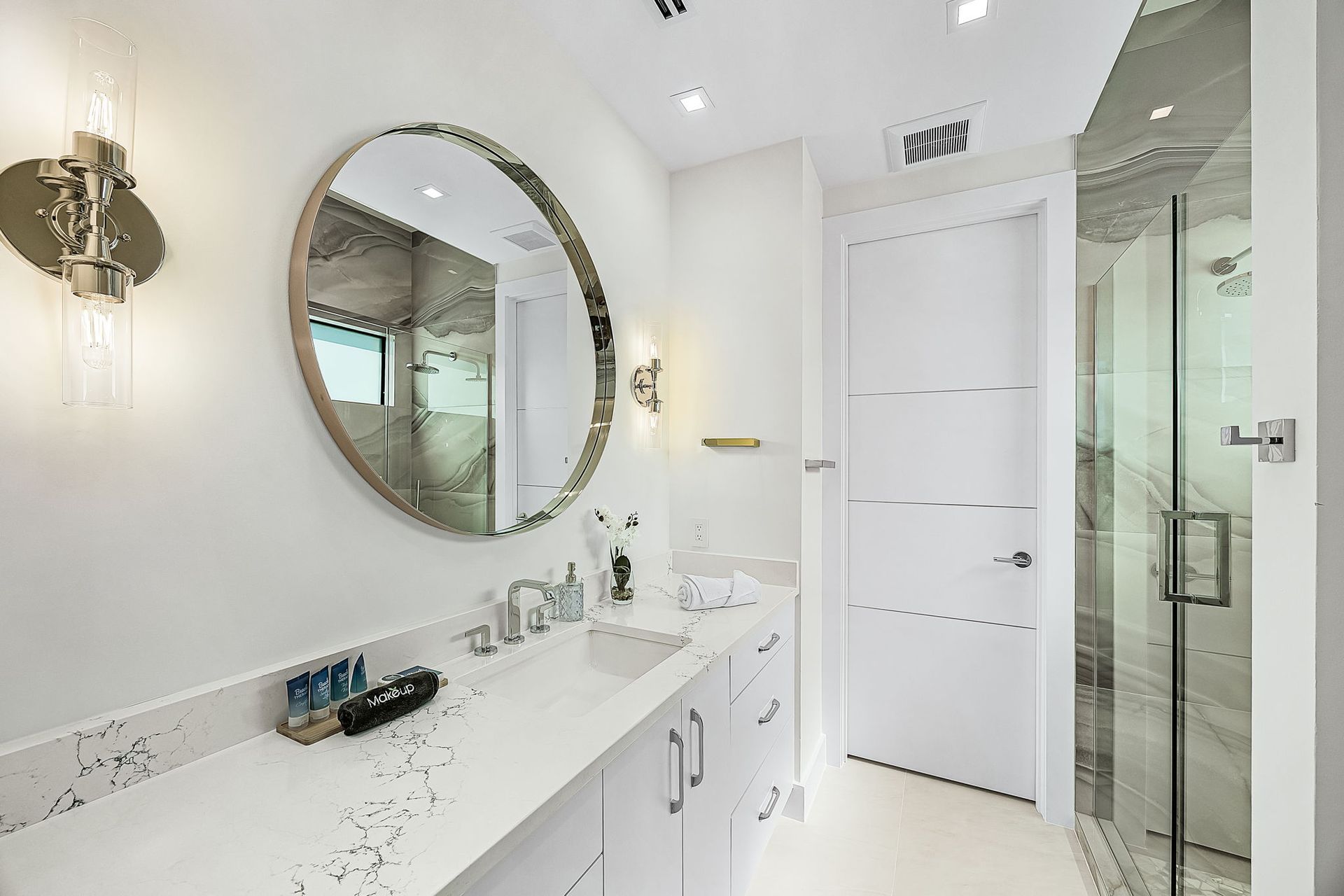 Modern white bathroom with a long countertop, round mirror, and glass shower.