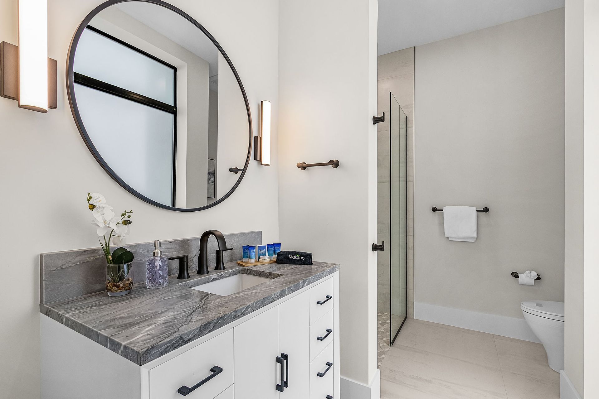 Bathroom with white cabinets, gray countertop, black fixtures, and a large round mirror.