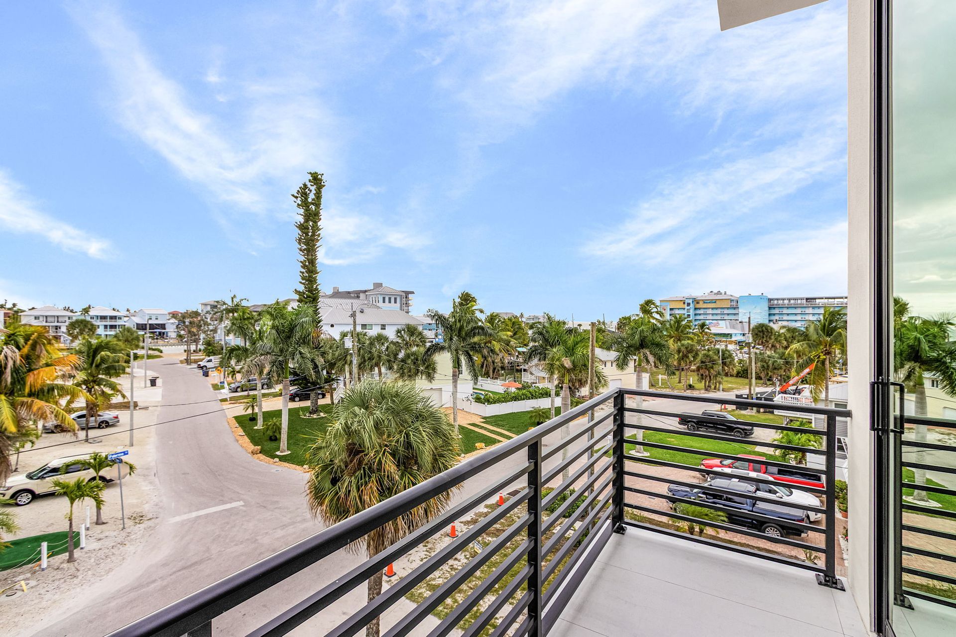 Balcony view of street lined with palm trees, buildings, and parked cars under a bright blue sky.