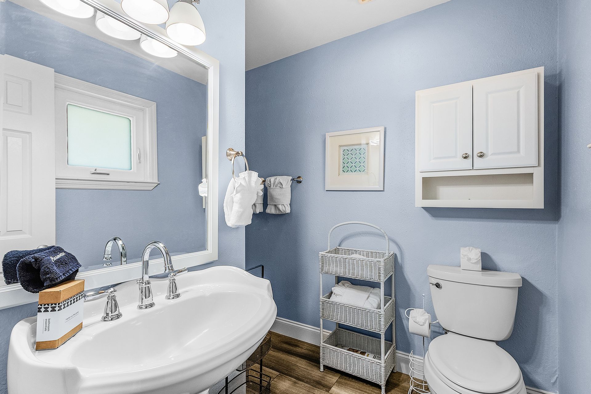 Bright blue bathroom with white vanity, toilet, wall cabinet, mirror, and metal storage rack.