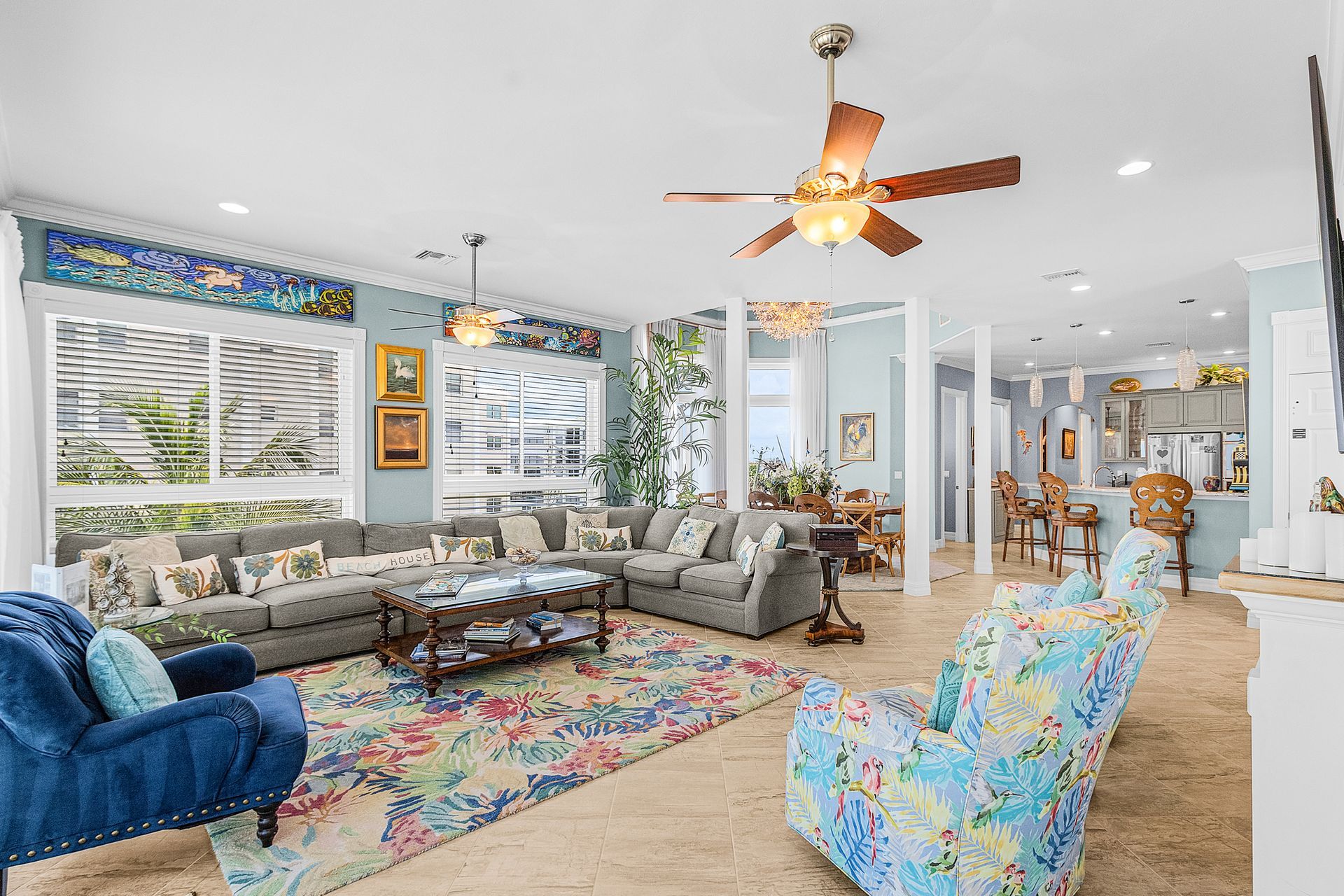 Bright open living room with colorful floral decor, gray sectional, blue chairs, and ceiling fan