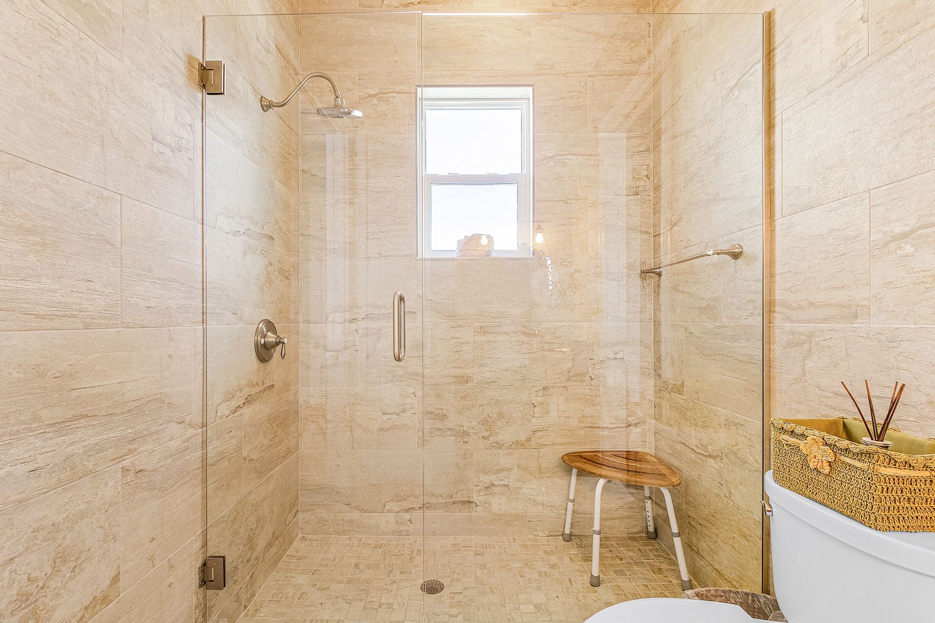 Beige tiled walk-in shower with glass door, window, and small stool beside a sink.