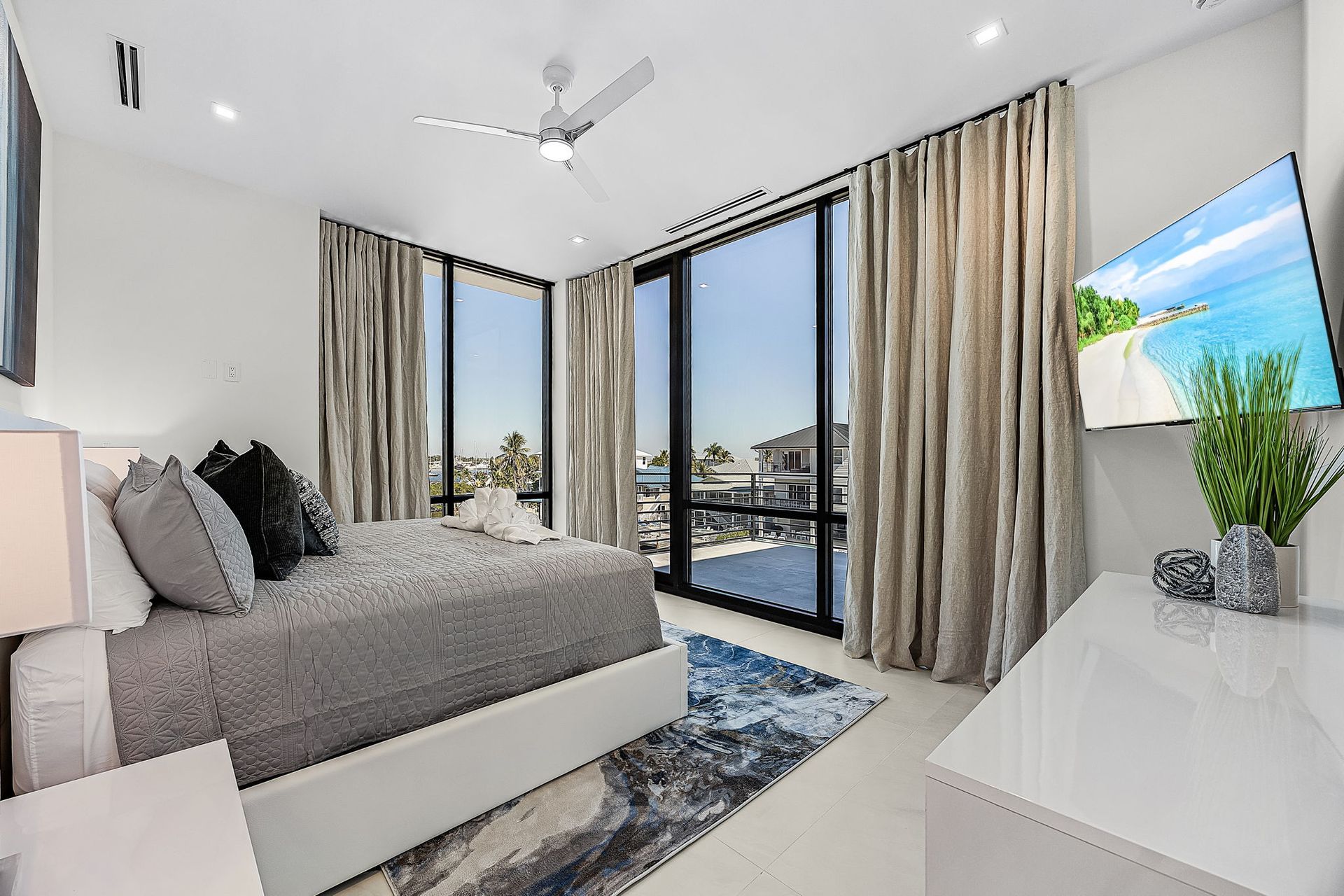 Bedroom with large windows, gray bed, white dresser, and beach scene on TV.