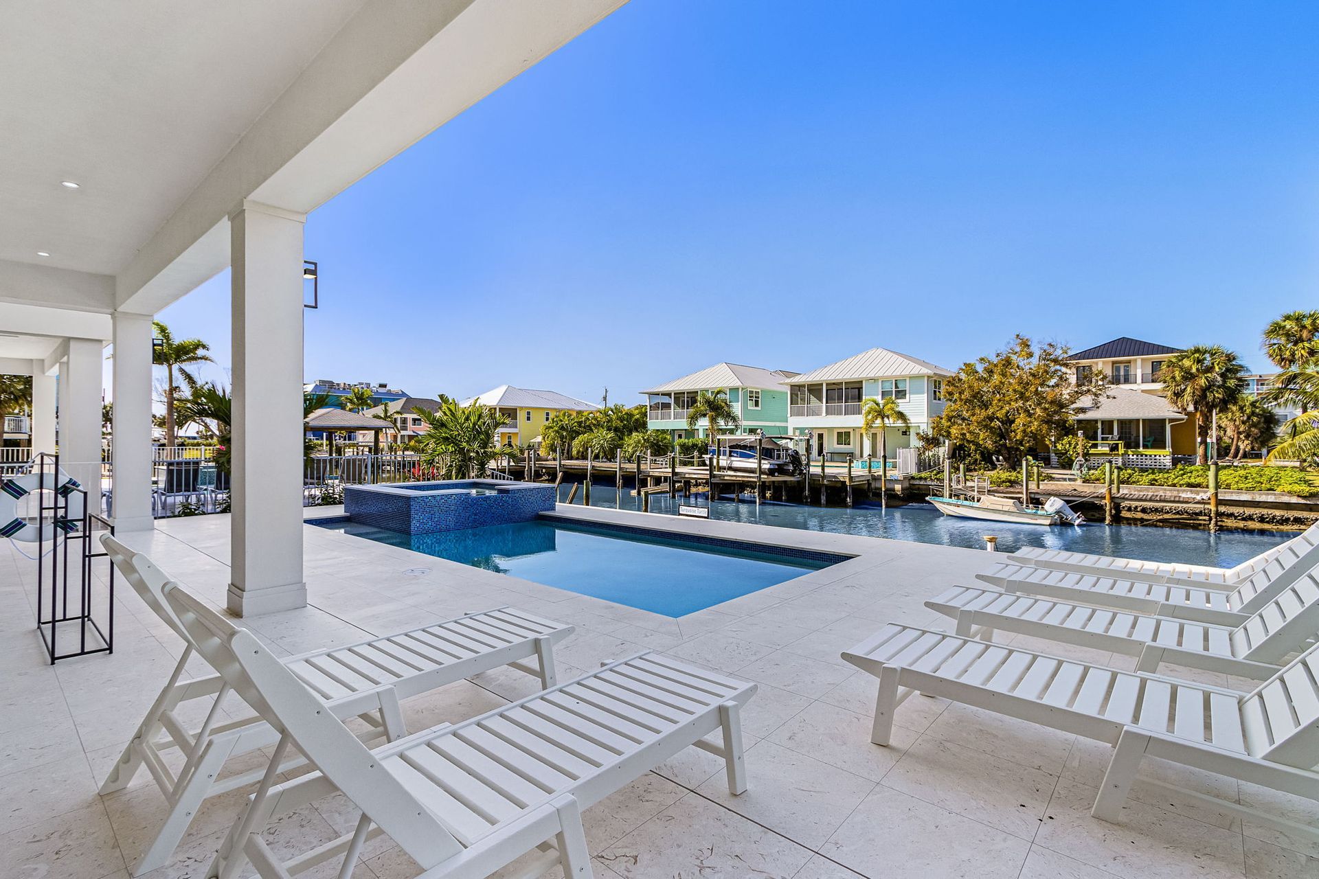 Patio with white lounge chairs overlooking a canal with a pool in the foreground and houses in the background.