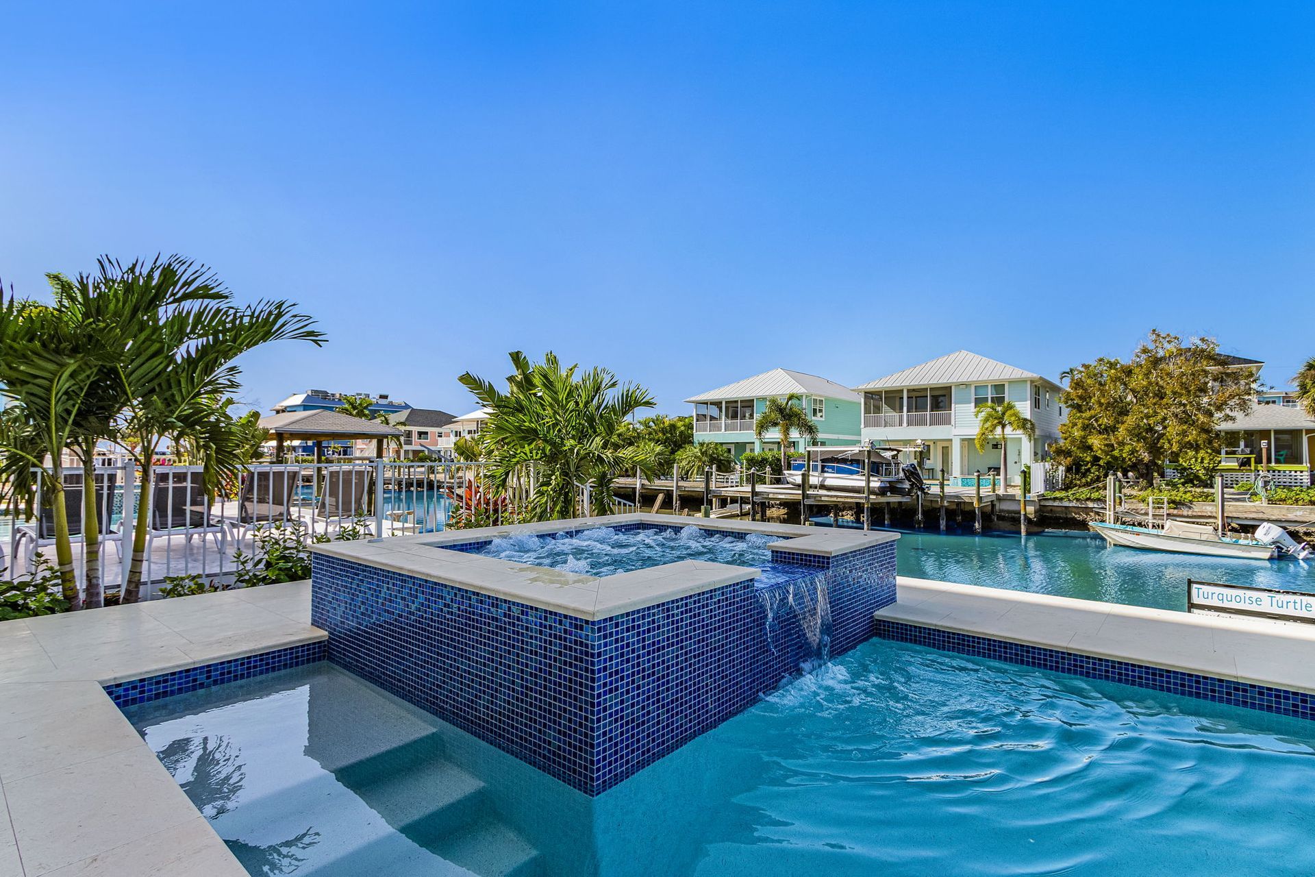 Pool with a blue tiled waterfall feature overlooking a canal, palm trees, and waterfront homes under a clear sky.