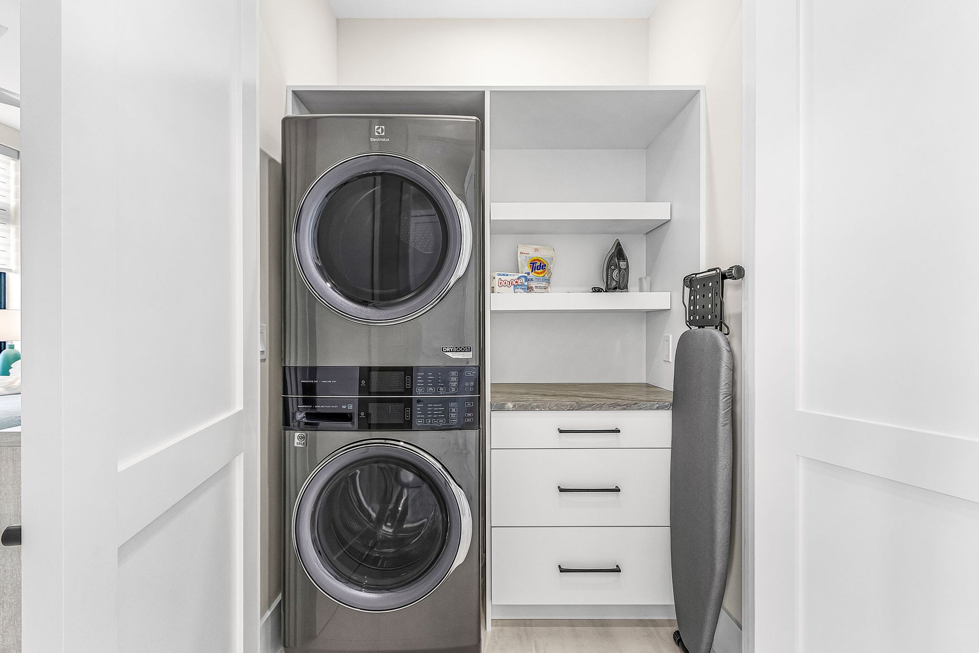 Stackable washer/dryer in a white laundry room with shelving, countertop, and ironing board.