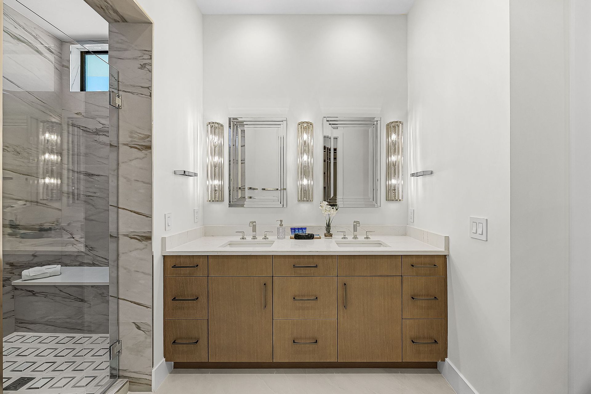 Bathroom with a double vanity, wood cabinets, mirrors with sconces, and a marble shower.