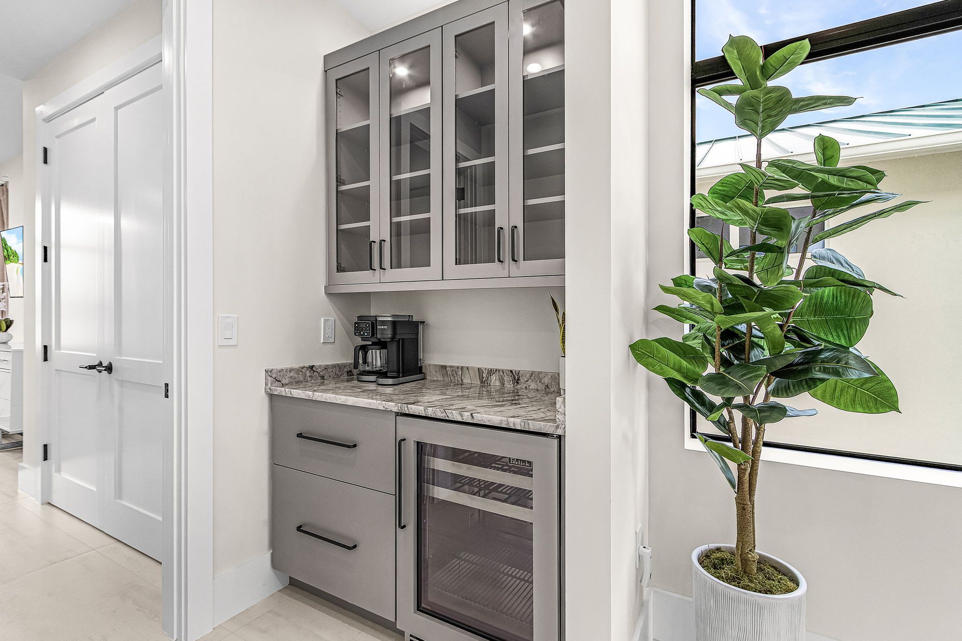 Built-in gray kitchen bar with cabinets, a small refrigerator, coffee maker, and a potted plant by a window.