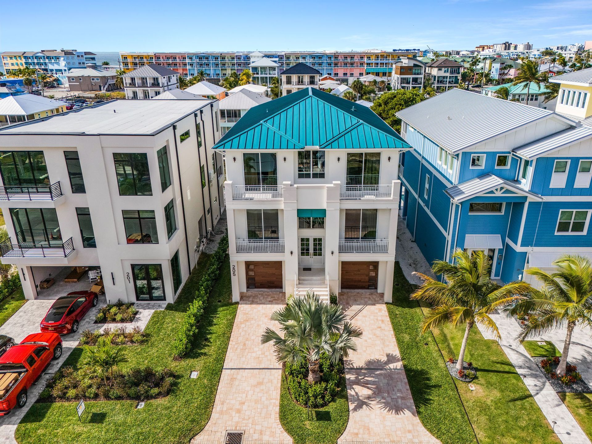 Multi-story white house with teal roof, flanked by homes, palm trees, and parked cars in a sunny neighborhood.